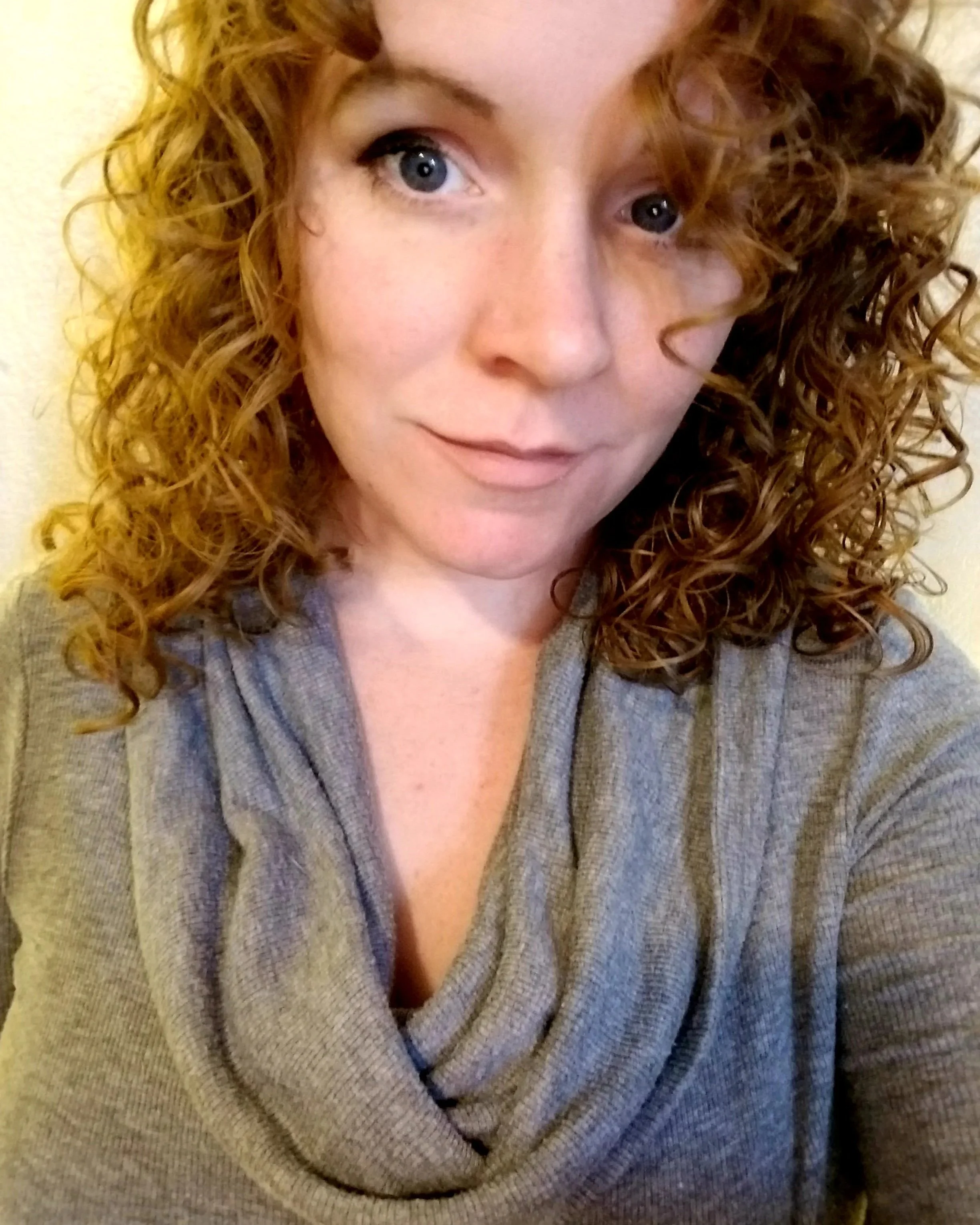 Close-up of a woman with curly red hair, blue eyes, light skin, and wearing a gray v-neck sweater.