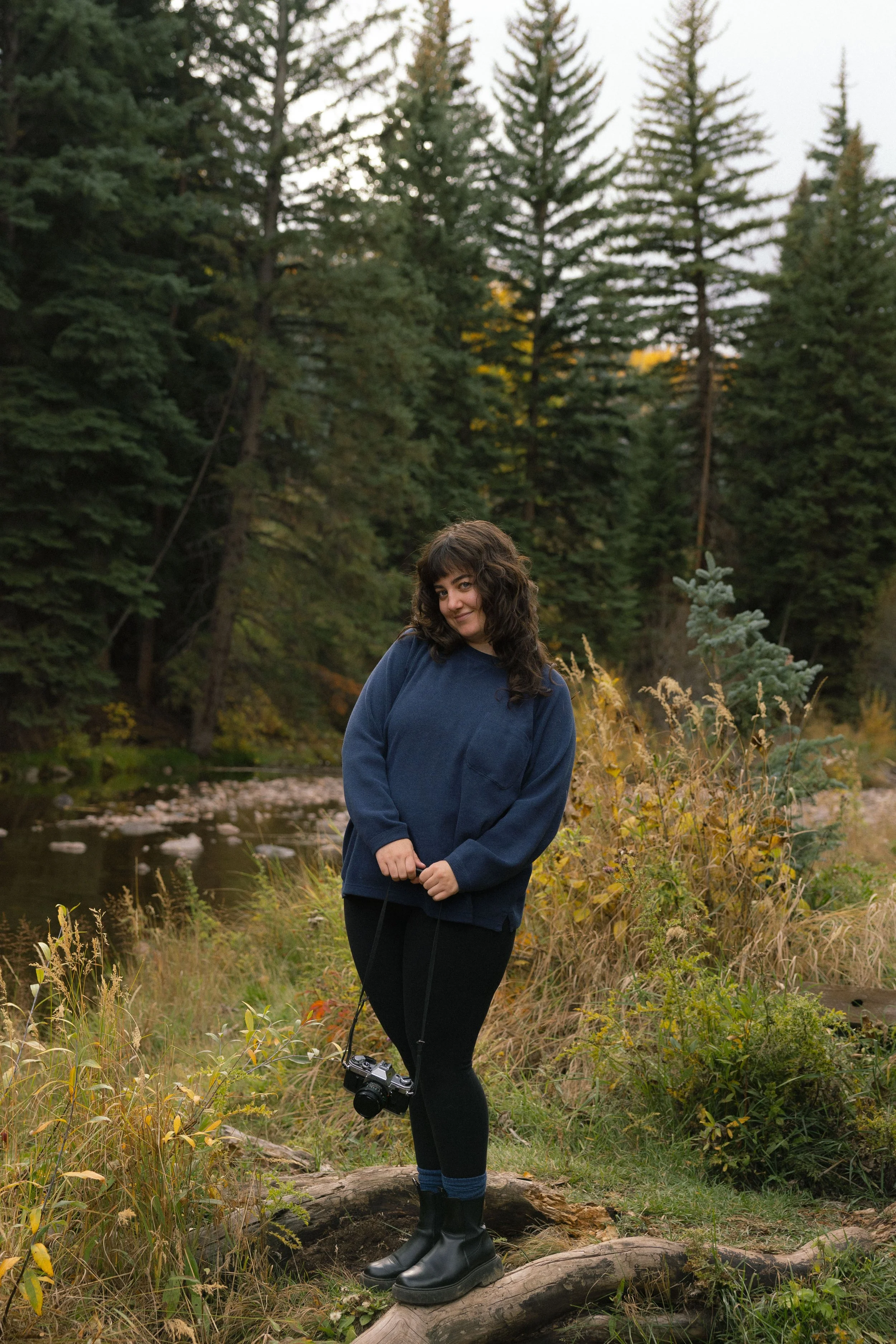 A woman with dark curly hair wearing a navy blue jacket, black leggings, and black boots standing on a fallen tree log near a river in a forested area during autumn. She is holding a camera and smiling at the camera with trees in the background.