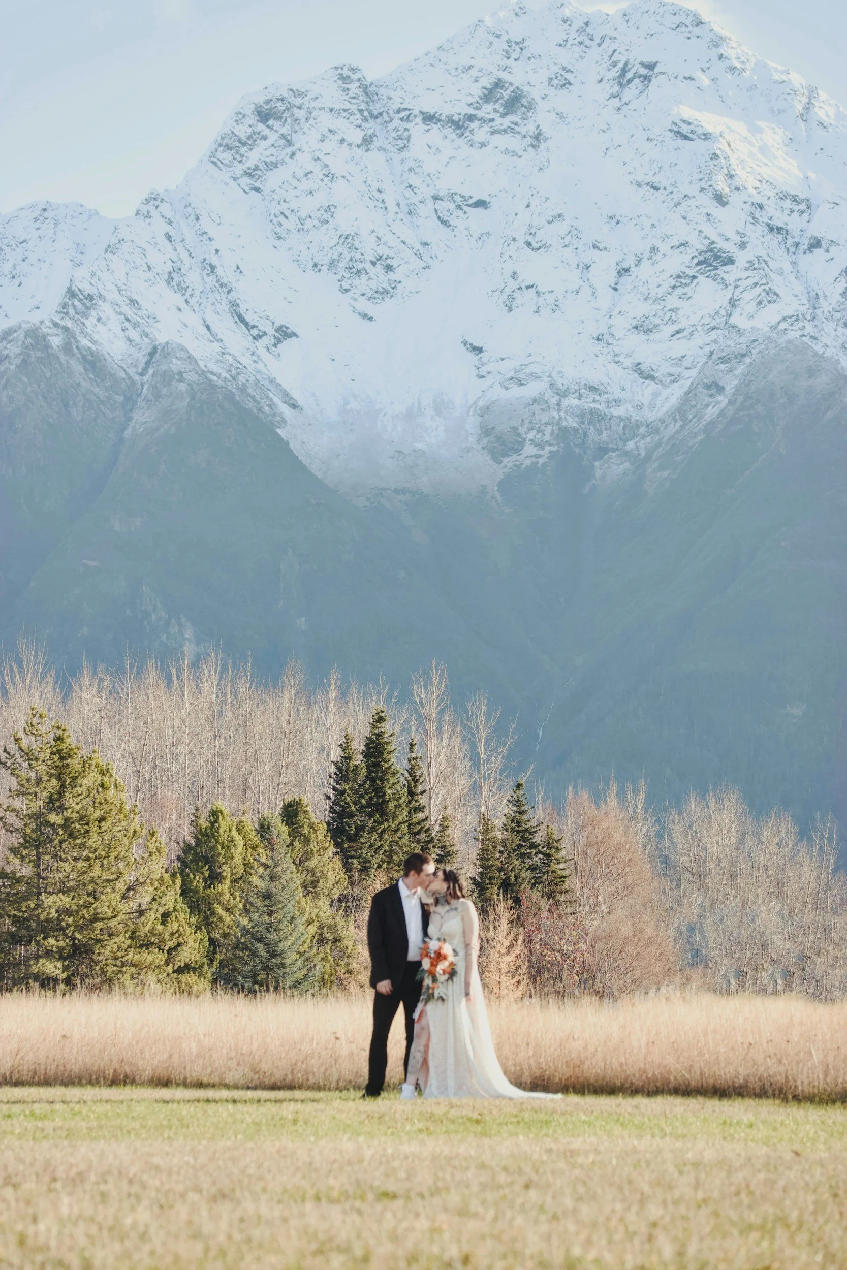 A bride and groom kissing outdoors with mountains in the background and trees surrounding them.