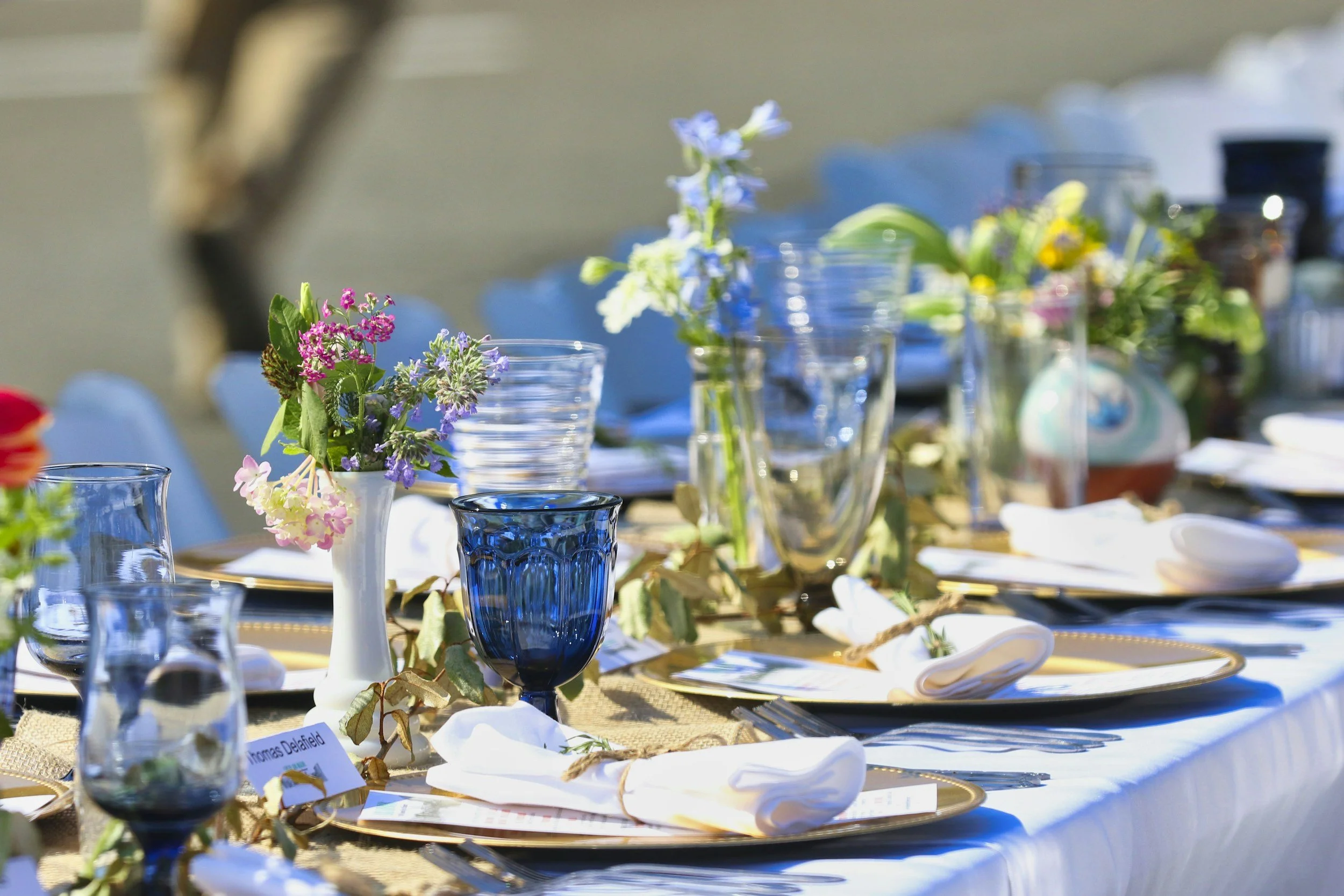 Table set for an outdoor event with floral centerpieces in glass vases and blue glasses, white plates, and napkins surrounded by greenery.