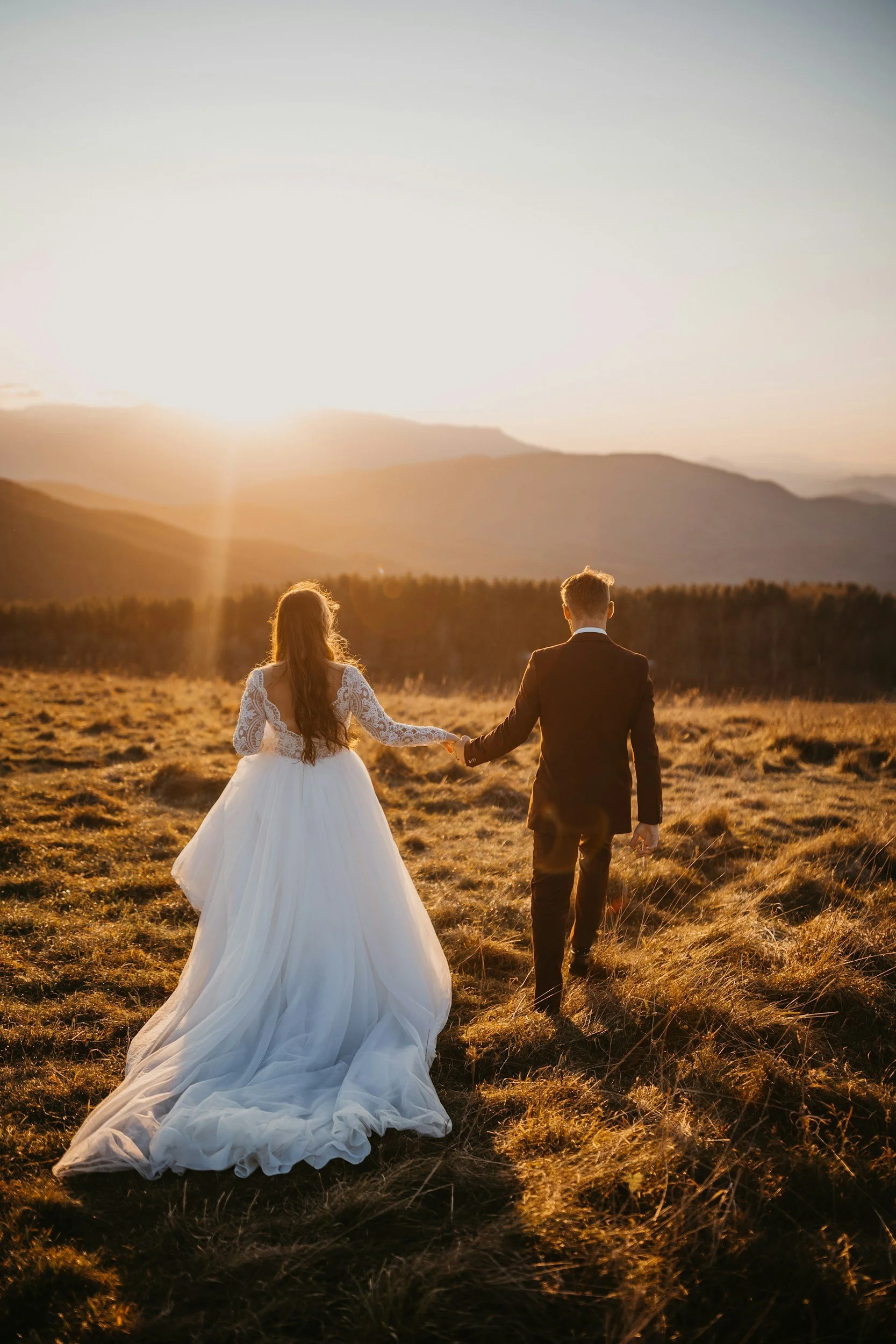 A bride and groom walking hand in hand through a grassy field during sunset with mountains in the background.
