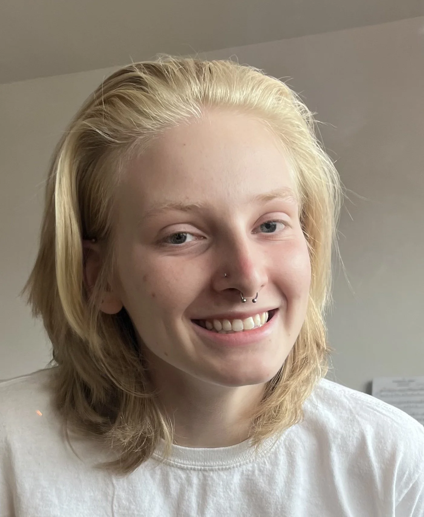 A young woman with blonde, shoulder-length hair, light skin, and multiple facial piercings smiling in front of a plain wall.
