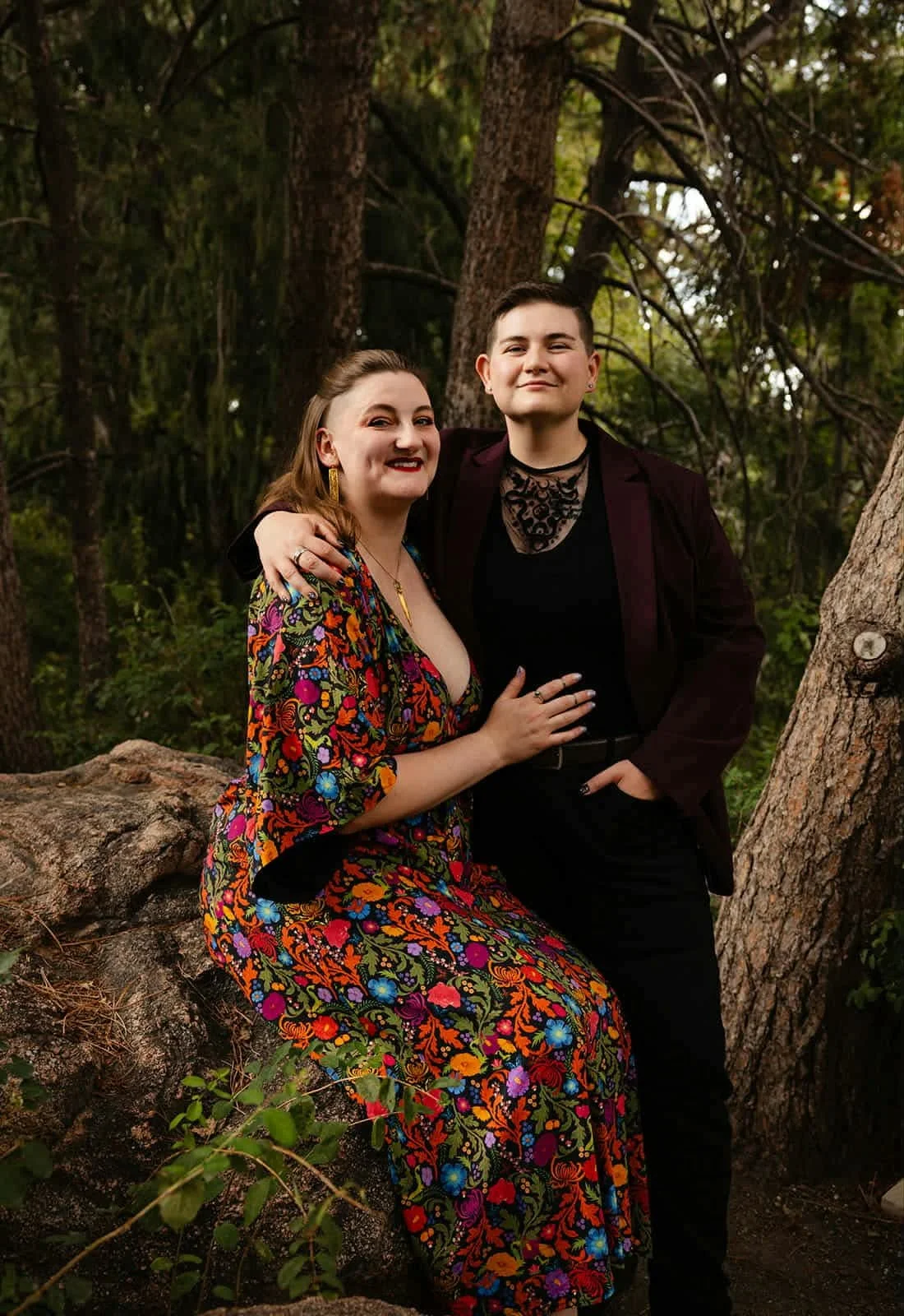 Two people pose for a photo outdoors in a wooded area, sitting on a large fallen tree trunk. One person is wearing a colorful floral dress and gold jewelry, while the other is in a dark outfit with a blazer and lace top. They are smiling and embracing each other.