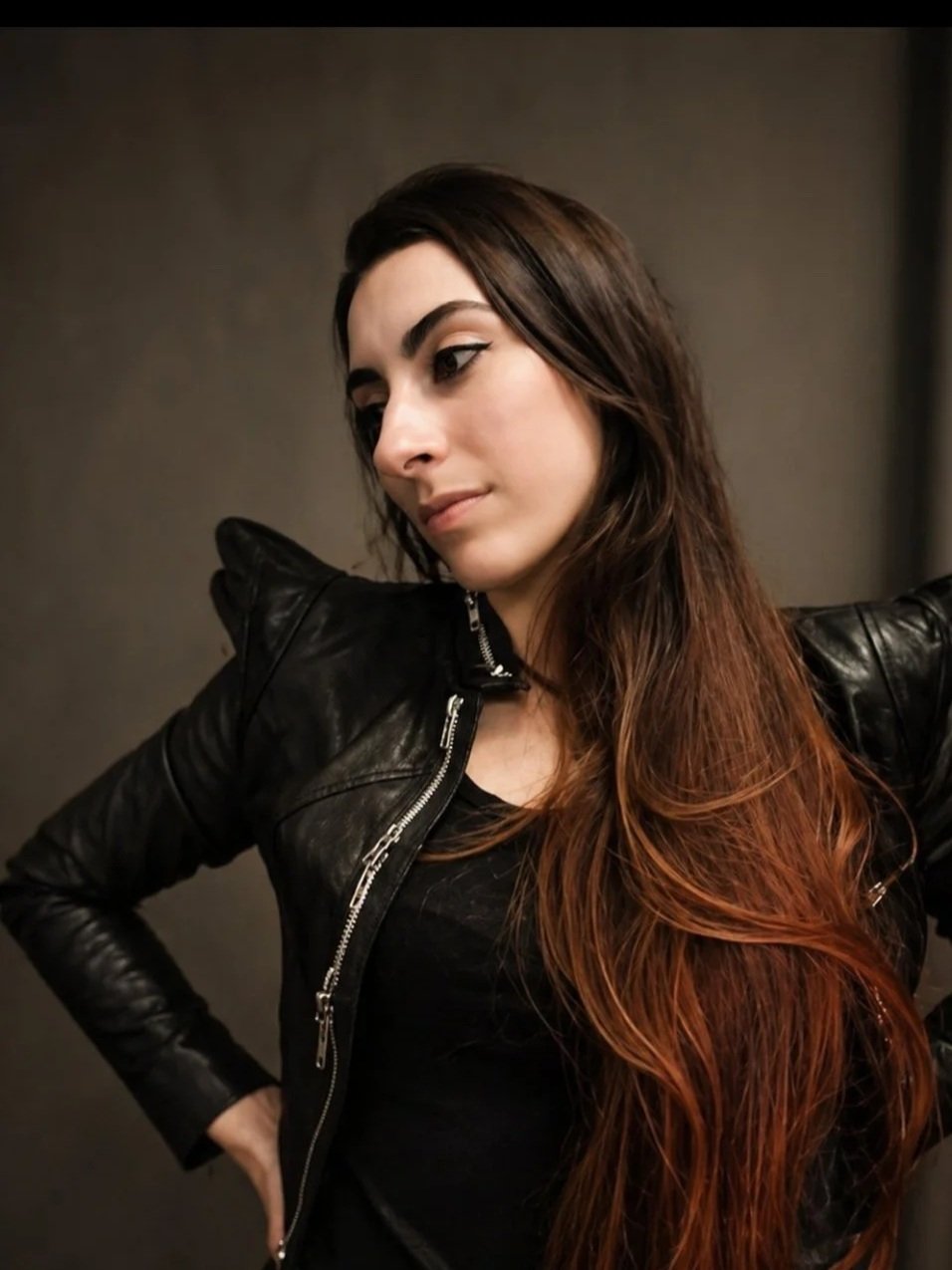 Portrait of a young woman with long, wavy hair wearing a black leather jacket, standing against a dark background.