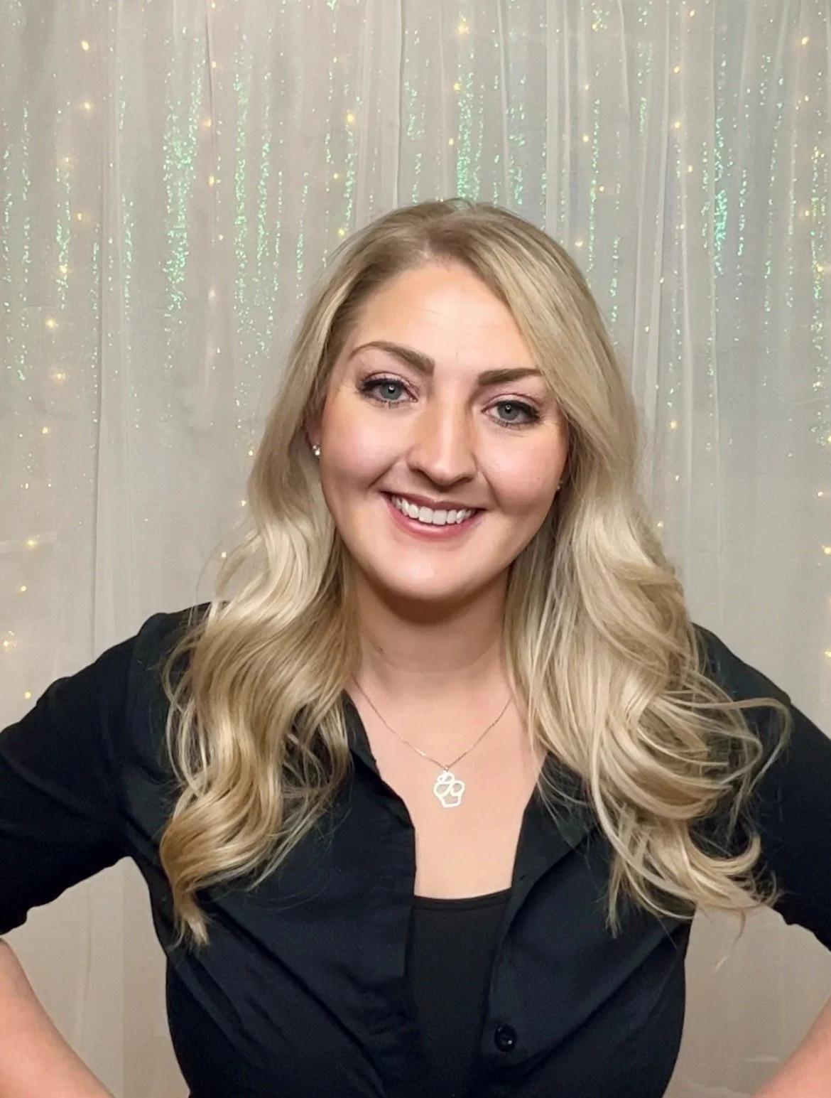 A smiling woman with long blonde hair, wearing a black shirt and a heart-shaped necklace, standing in front of a background with string lights.