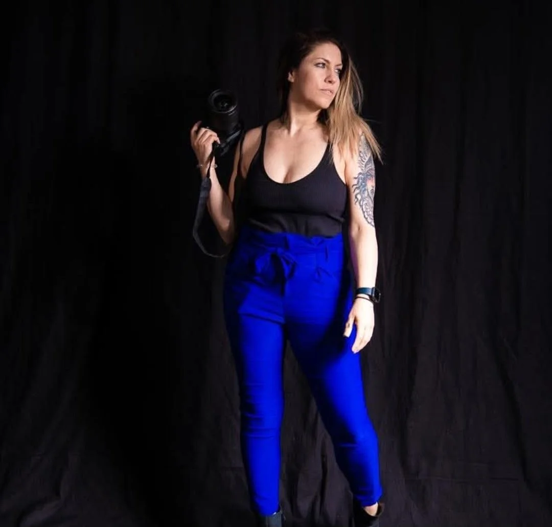 A woman with long hair and a tattoo on her arm, dressed in a black top and blue pants, standing against a black backdrop, holding a camera over her shoulder and looking to the side.