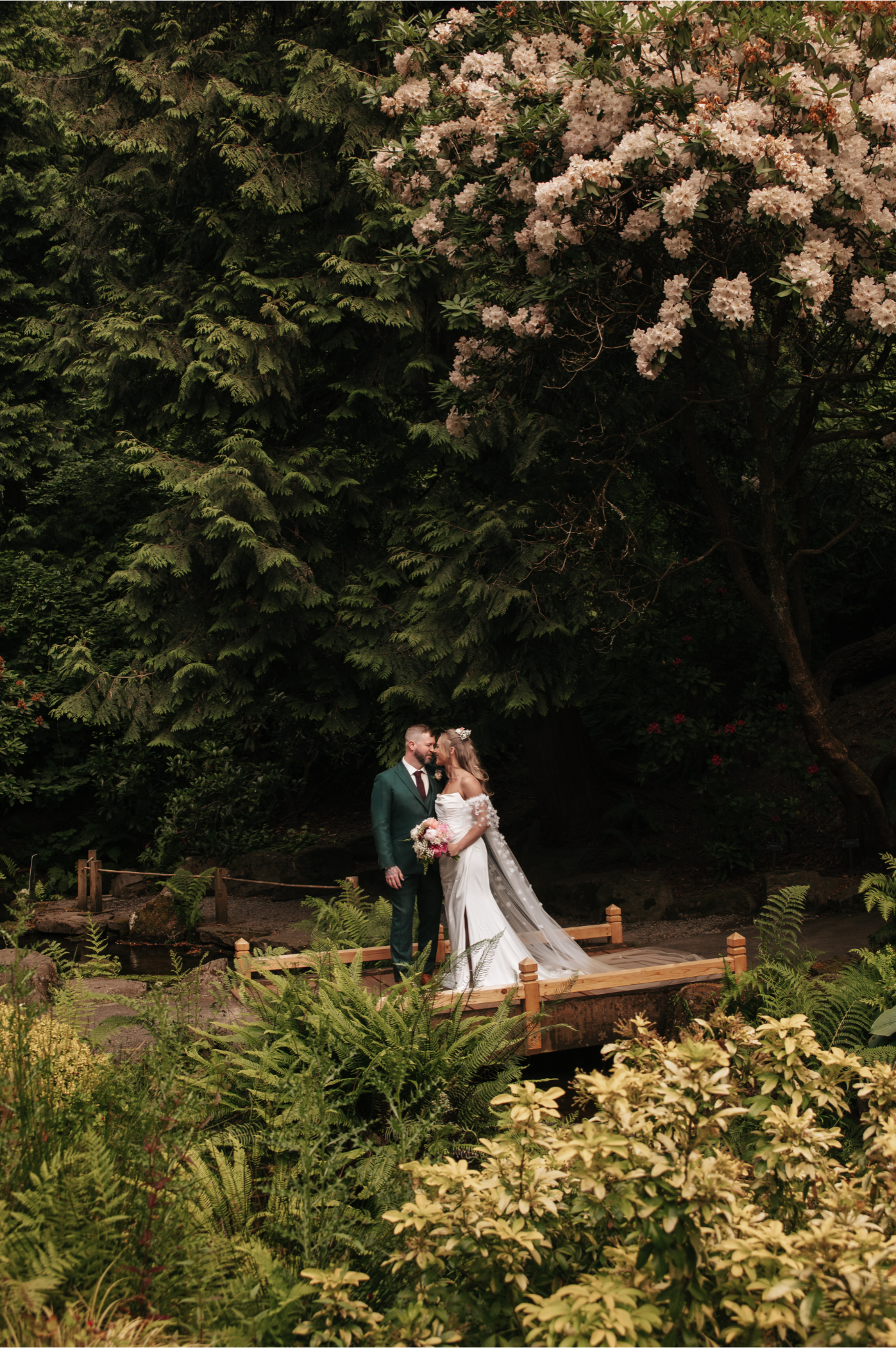 A bride and groom stand on a small wooden bridge surrounded by lush greenery and flowering plants, with large trees and blooming pink flowers in the background.