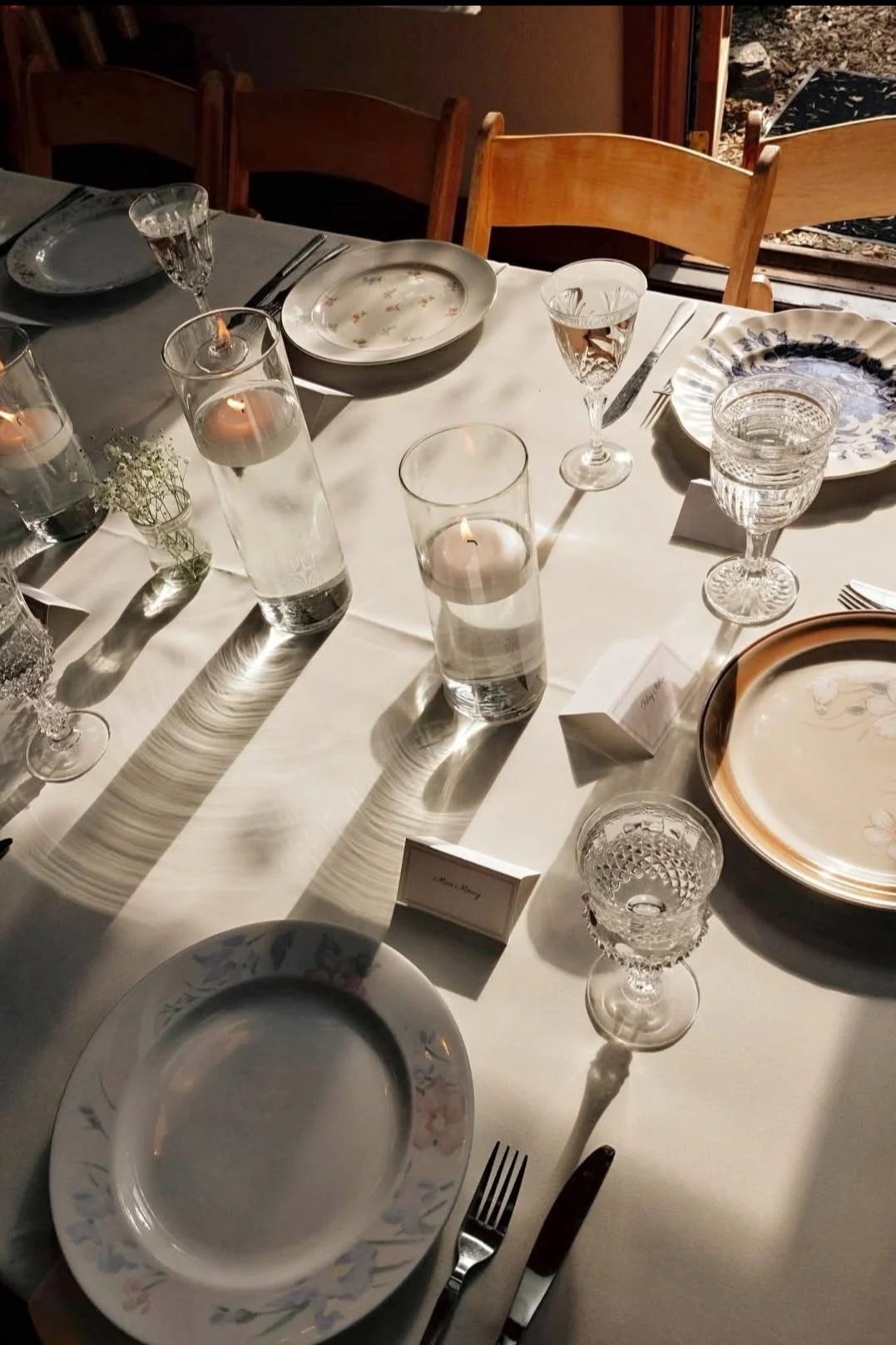 A formal dining table set with plates, glasses, candles, and utensils for a meal, with a white tablecloth and wooden chairs.