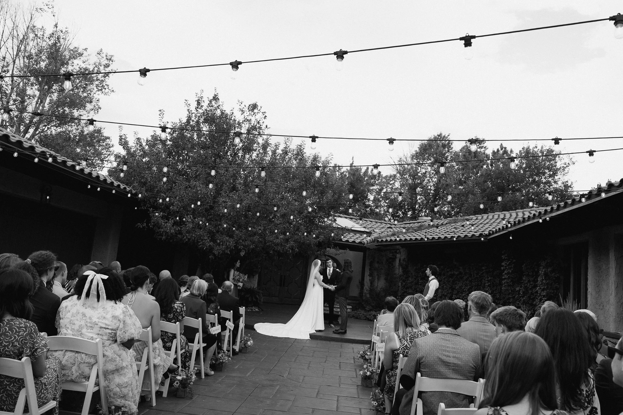 Wedding ceremony outdoors with guests seated under string lights, bride and groom holding hands in front of officiant, with trees and a tiled roof in the background.