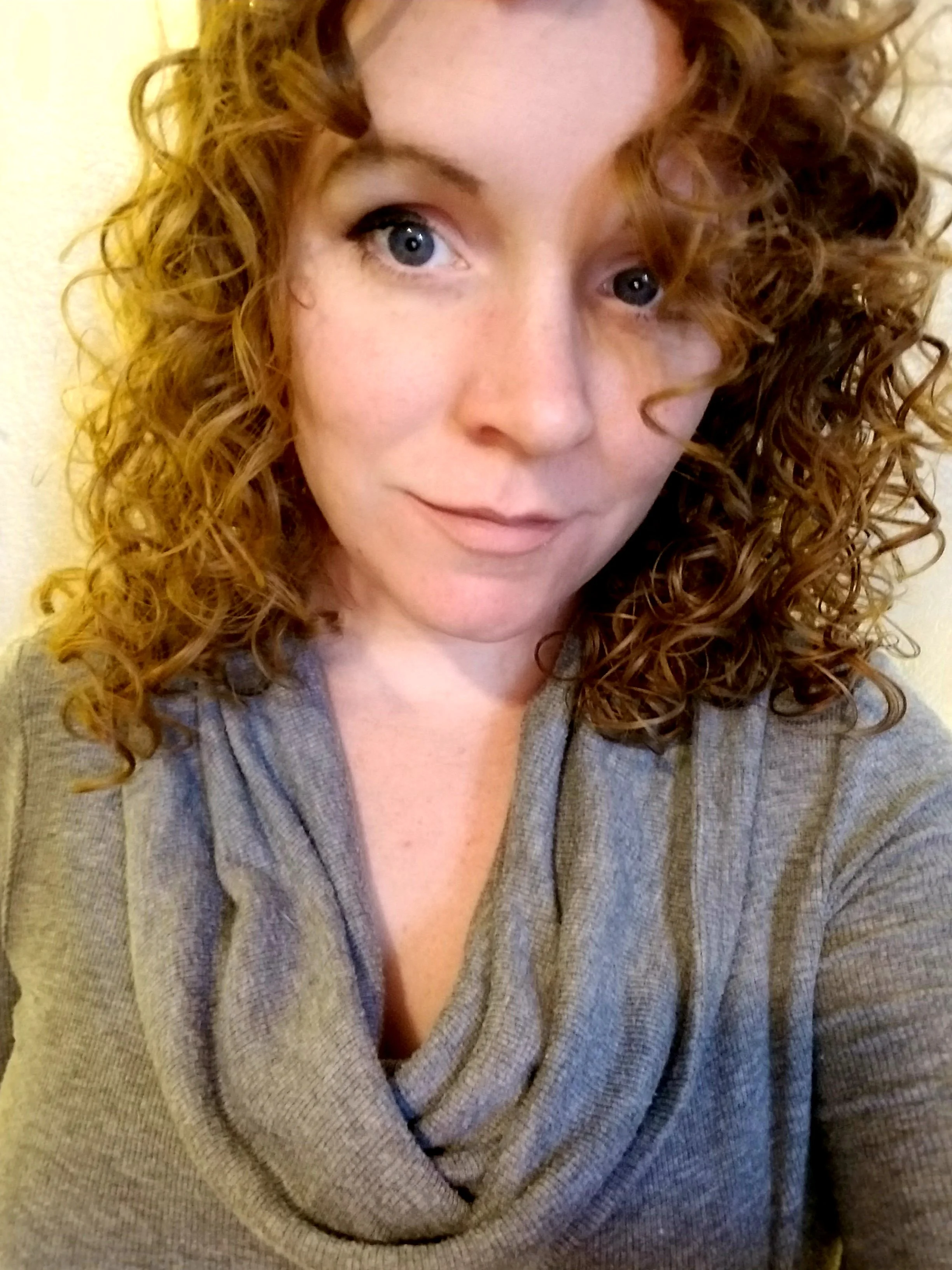 A woman with curly red hair and blue eyes, wearing a gray sweater, looking off to the side.