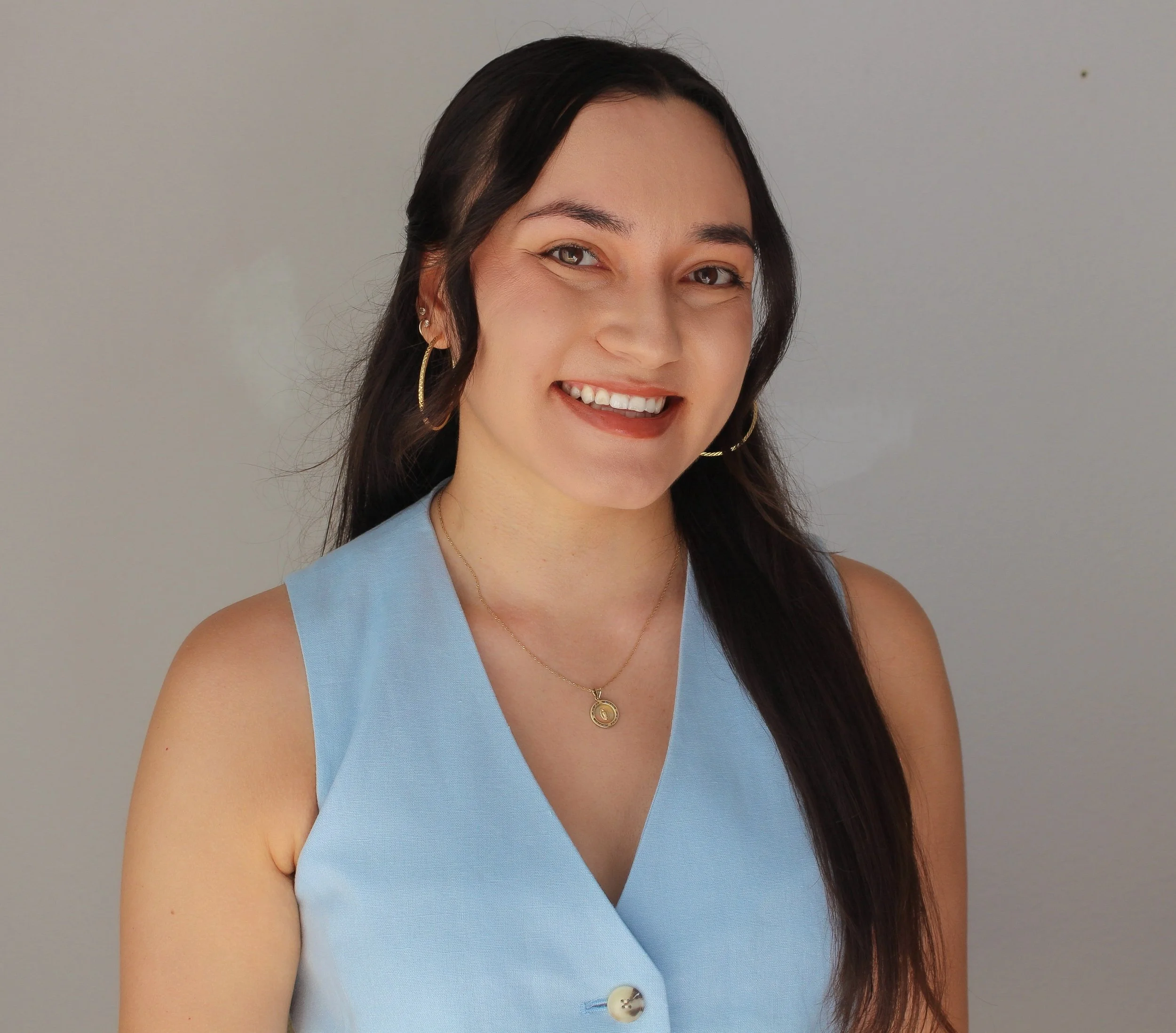 A woman with long dark hair, wearing a light blue sleeveless top, gold hoop earrings, and a gold necklace, smiling against a plain light gray background.