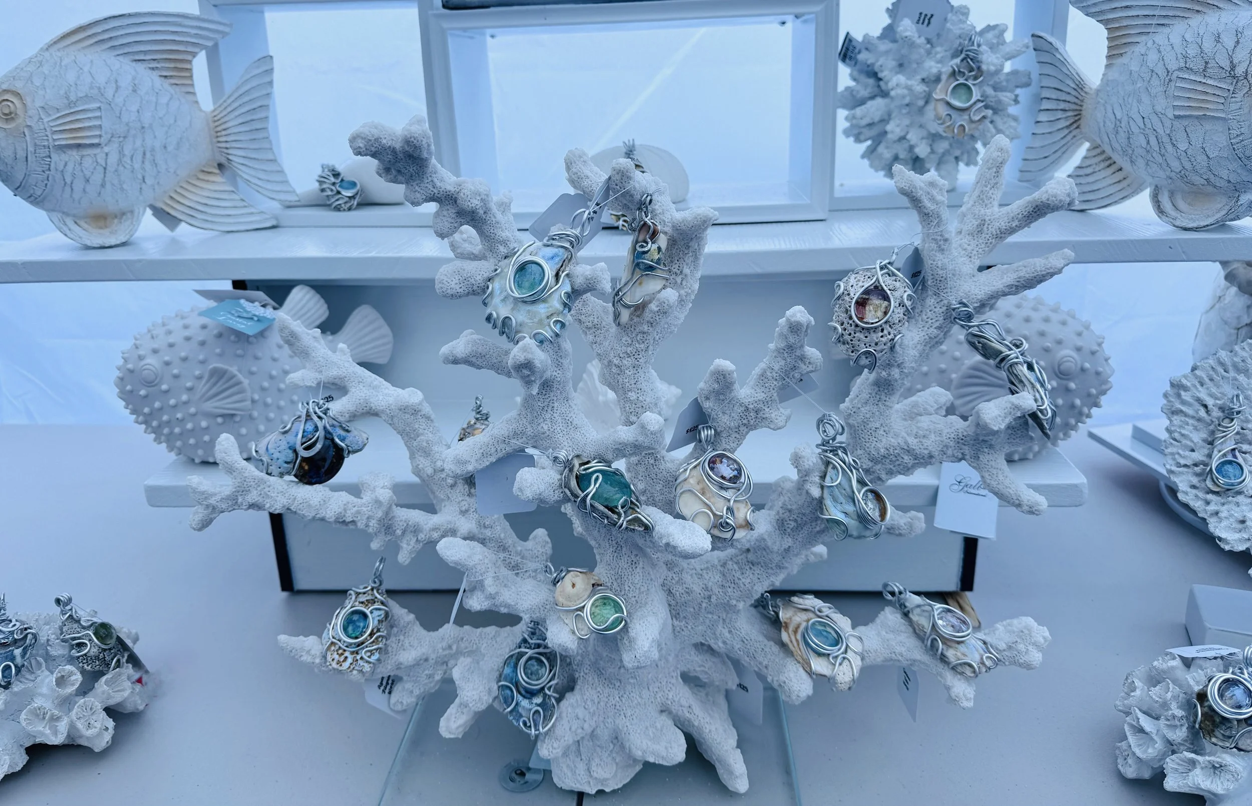 Display of jewelry including rings and necklaces on white coral branches with white fish sculptures in the background, inside a transparent case.