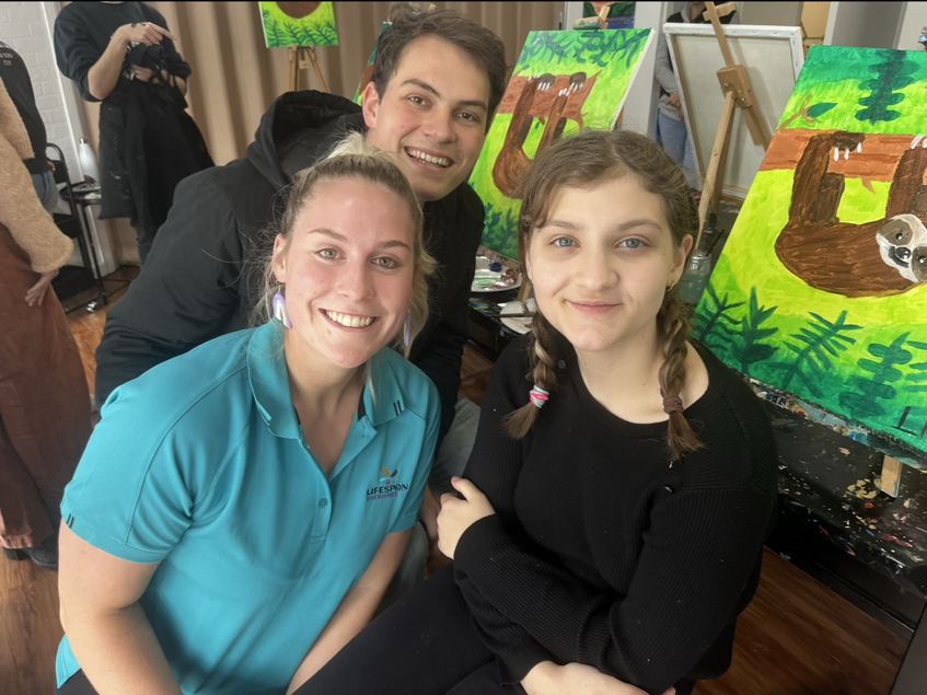 Occupational Therapist Georgia Black and occupational therapist Ryan smiling with a child, with two painted canvases of sloths on easels behind them in a classroom or art studio setting.