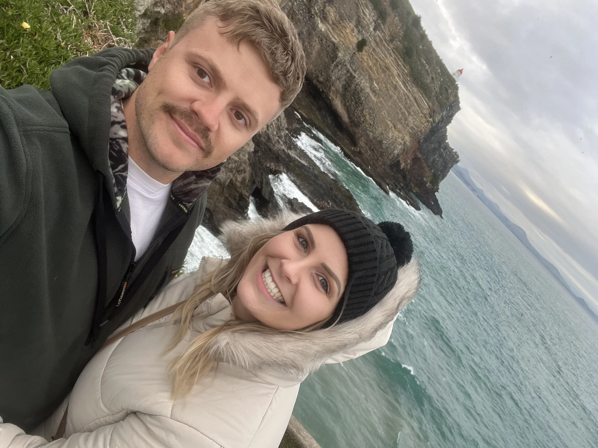A smiling couple taking a selfie at a rocky coastline, with the ocean and cliffs in the background on a cloudy day.