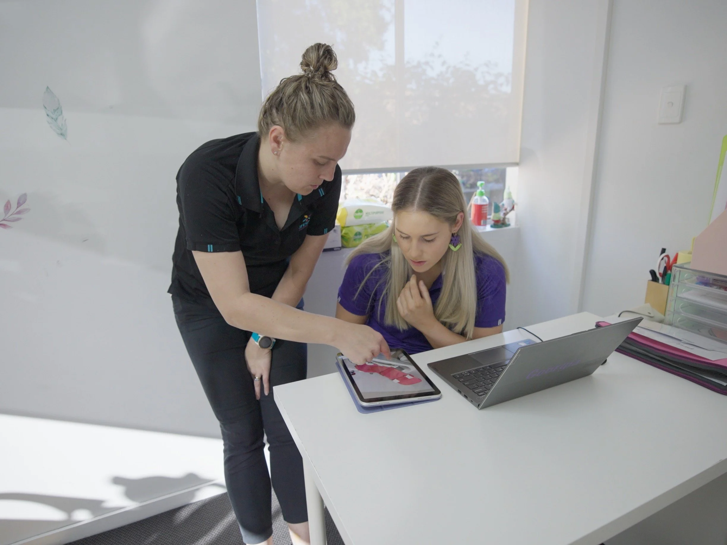 Two people in a Lifespan Therapies treatment room, physiotherapist Liz is standing and pointing at a tablet, while Georgia sitting and looking at it. Showcasing the career progression, mentoring and support available as a new graduate.