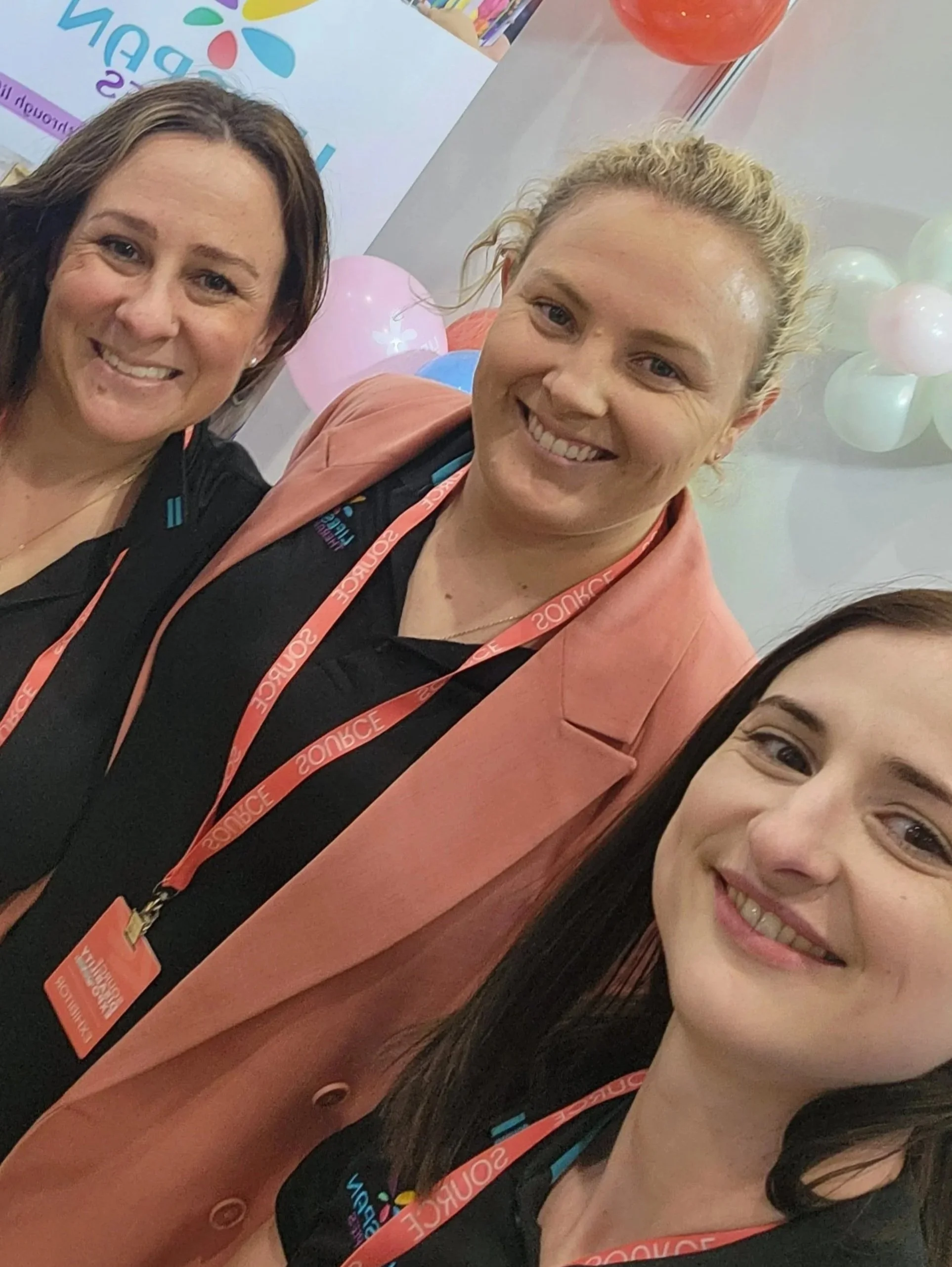 Three women smiling at a conference, wearing conference badges on red lanyards, with balloons and a colorful backdrop in the background.