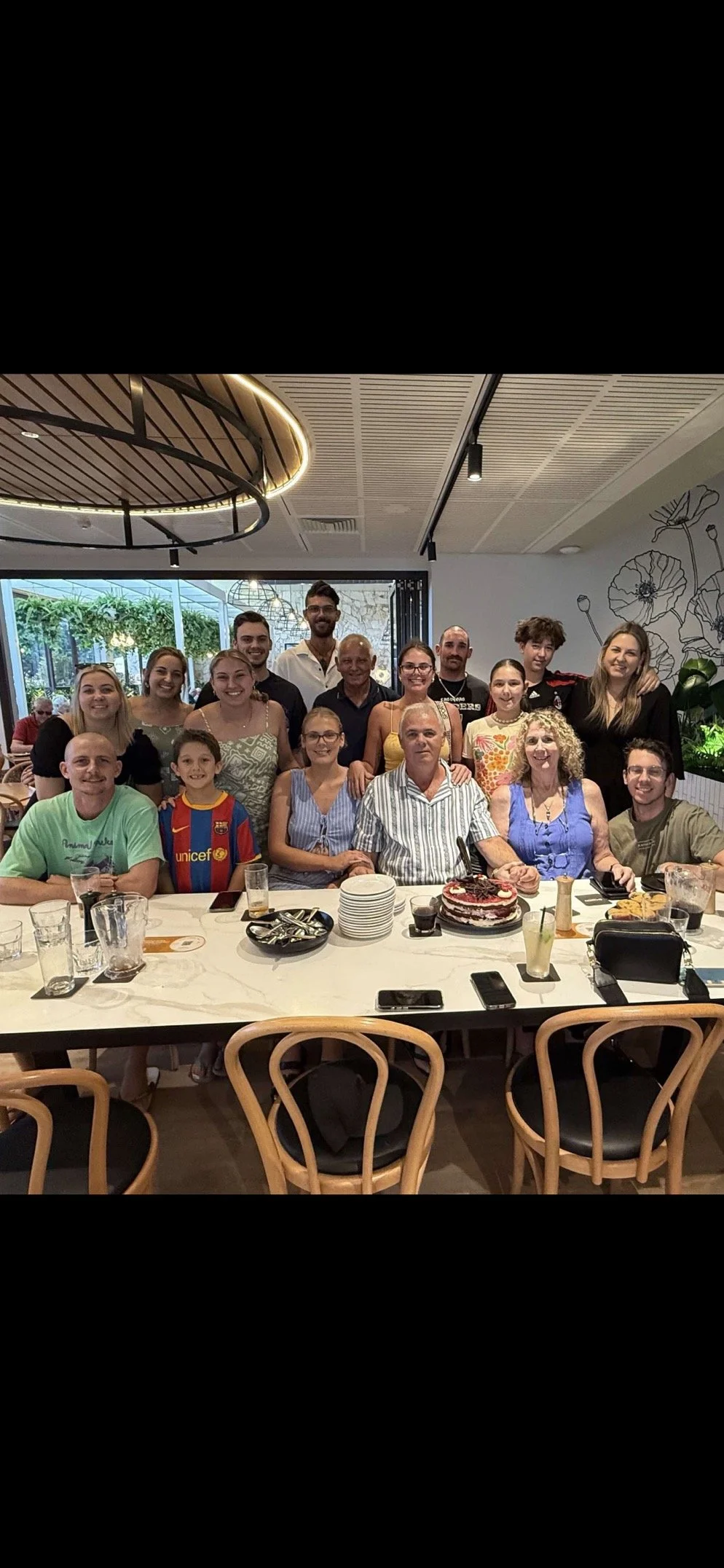 A group of people, including men, women, and children, are posing for a photo at a restaurant. They are gathered around a table with glasses, plates, and a dessert. The setting features modern decor with a ceiling light and a mural on the wall. The group looks happy and relaxed, suggesting a celebration or gathering.