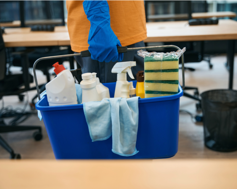 Person wearing orange shirt and blue gloves holding a blue cleaning caddy with various cleaning supplies, including spray bottles, a sponge, a cloth, and cleaning solutions, in an office setting.