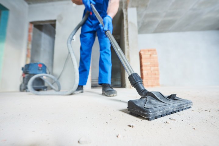 Worker in blue pants and gloves using a floor cleaning machine on a concrete surface under construction.