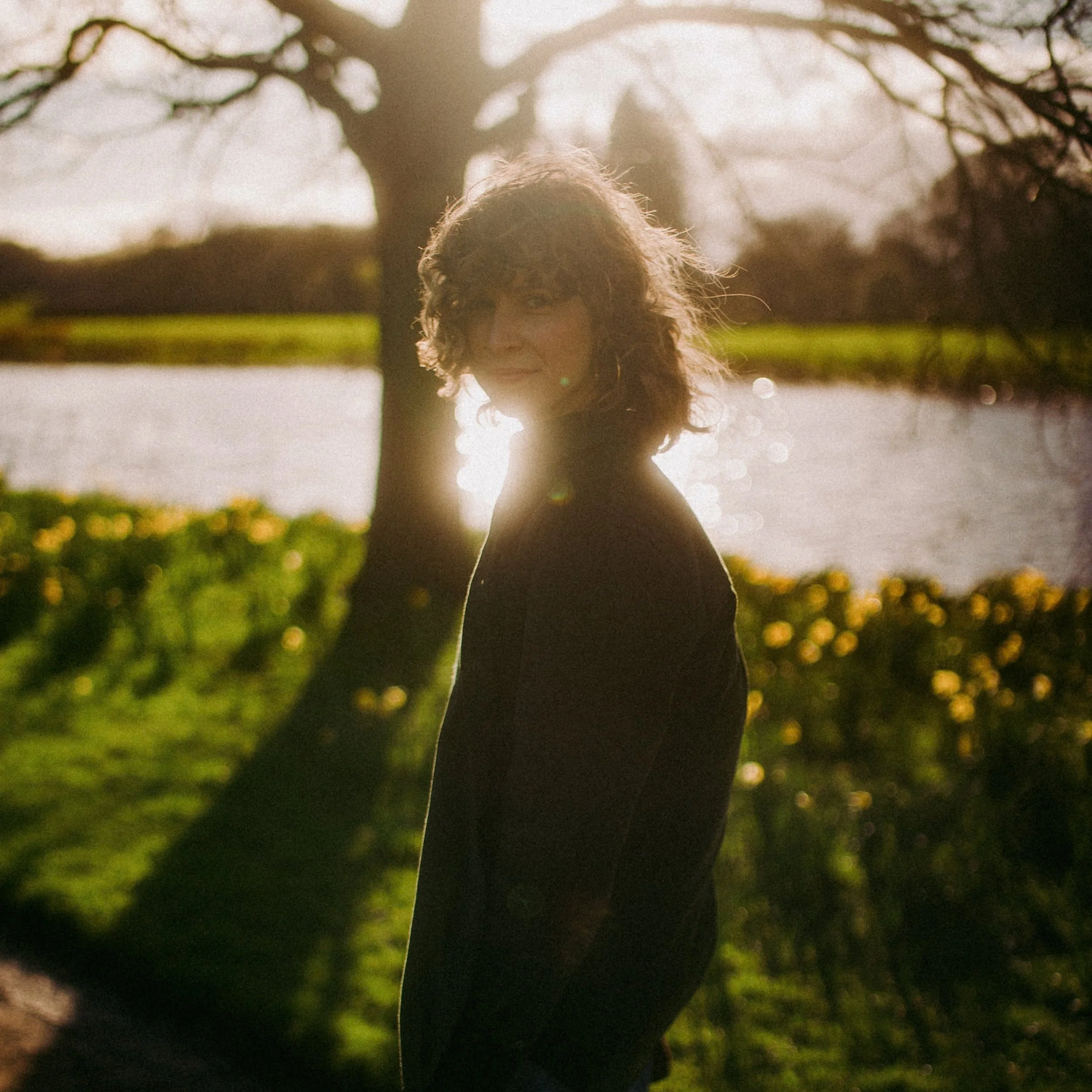 An artsy shot of Alisha strolling by a lake. The sun breaks between bare tree branches, so her face is backlit. She smiles at the camera and there are yellow out-of-focus flowers in the background.