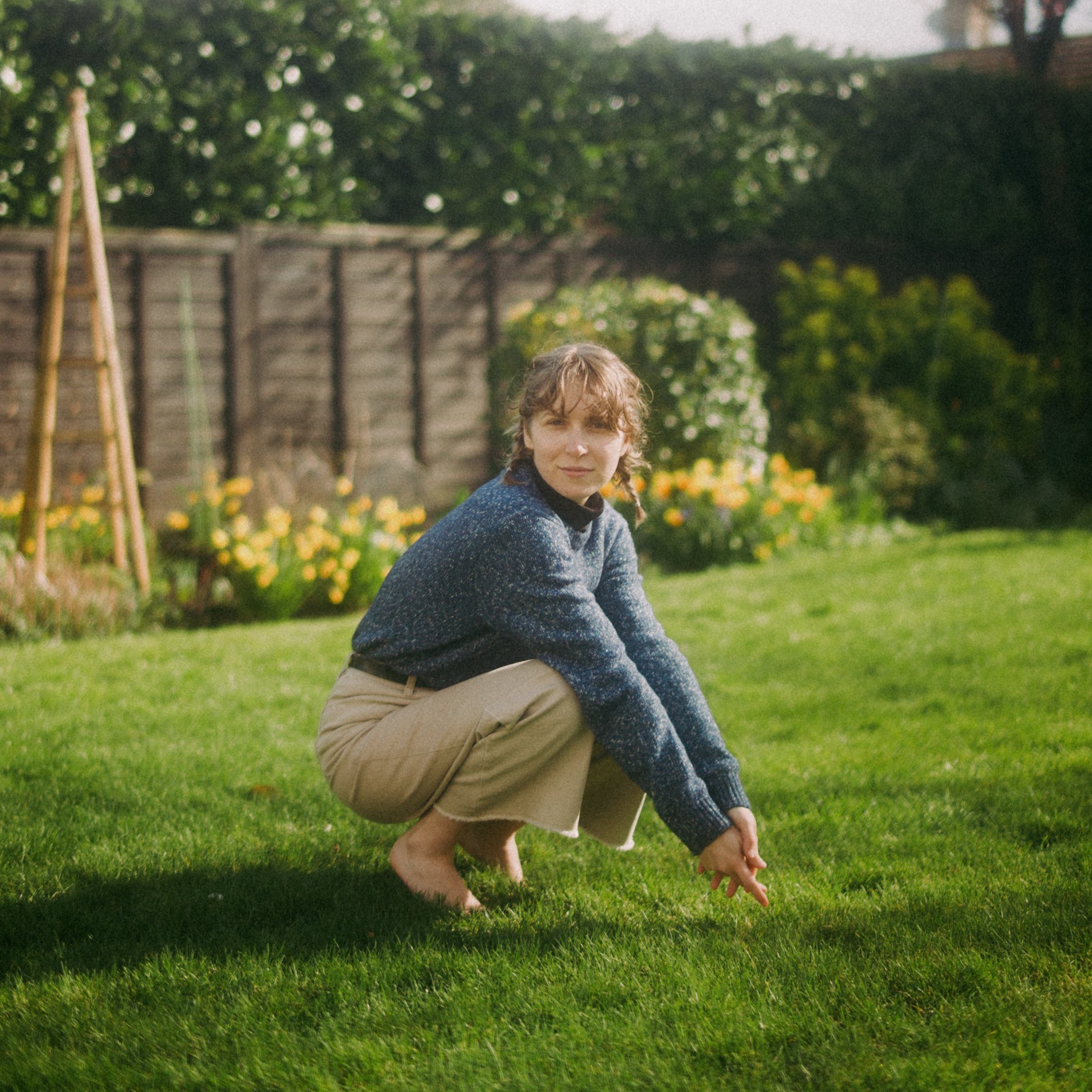 Alisha is squatting on a lush, green lawn. She wears a blue-grey sweater, wide-leg beige pants, and her hair is in braids. She looks straight at the camera, and there are yellow flowers blooming in the background.