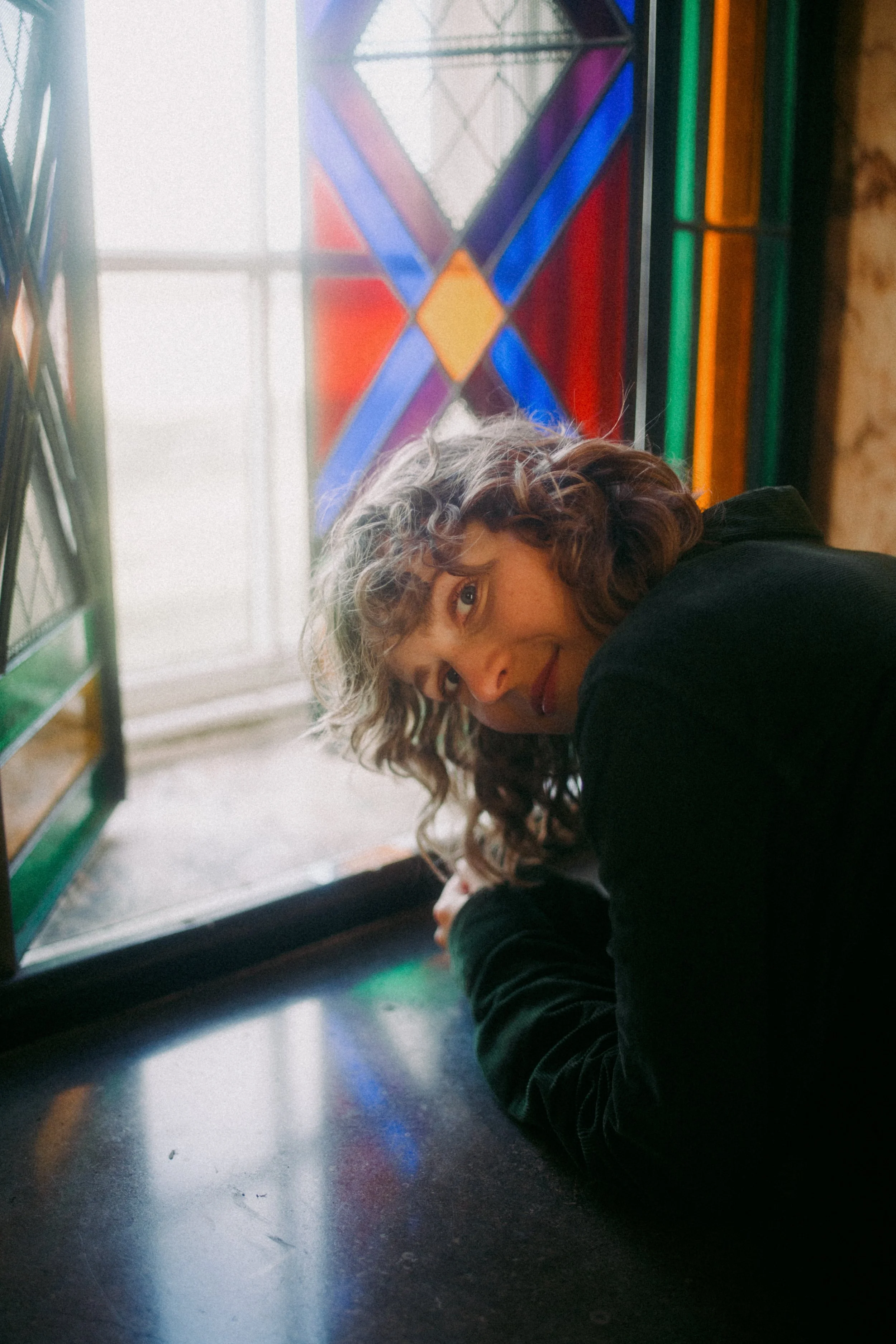 Alisha leans on a windowsill in front of stained glass. She is leaning forward and peeks up at the camera mischievously.