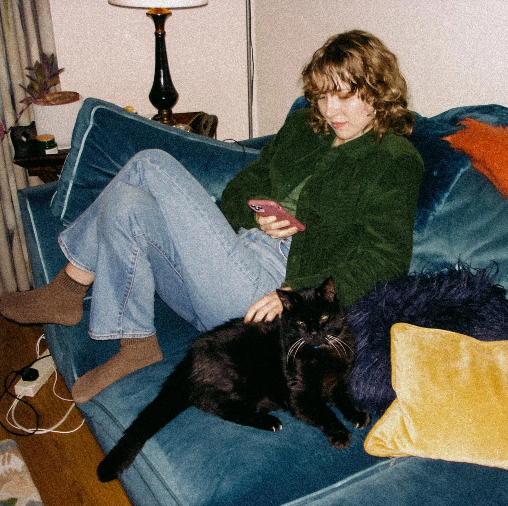 A film photograph of Alisha. She is wearing a green corduroy shirt and blue jeans. She sits on a blue velvet couch covered in cushions, patting a black cat while scrolling on her phone. The photograph is busy, colourful, and cosy.
