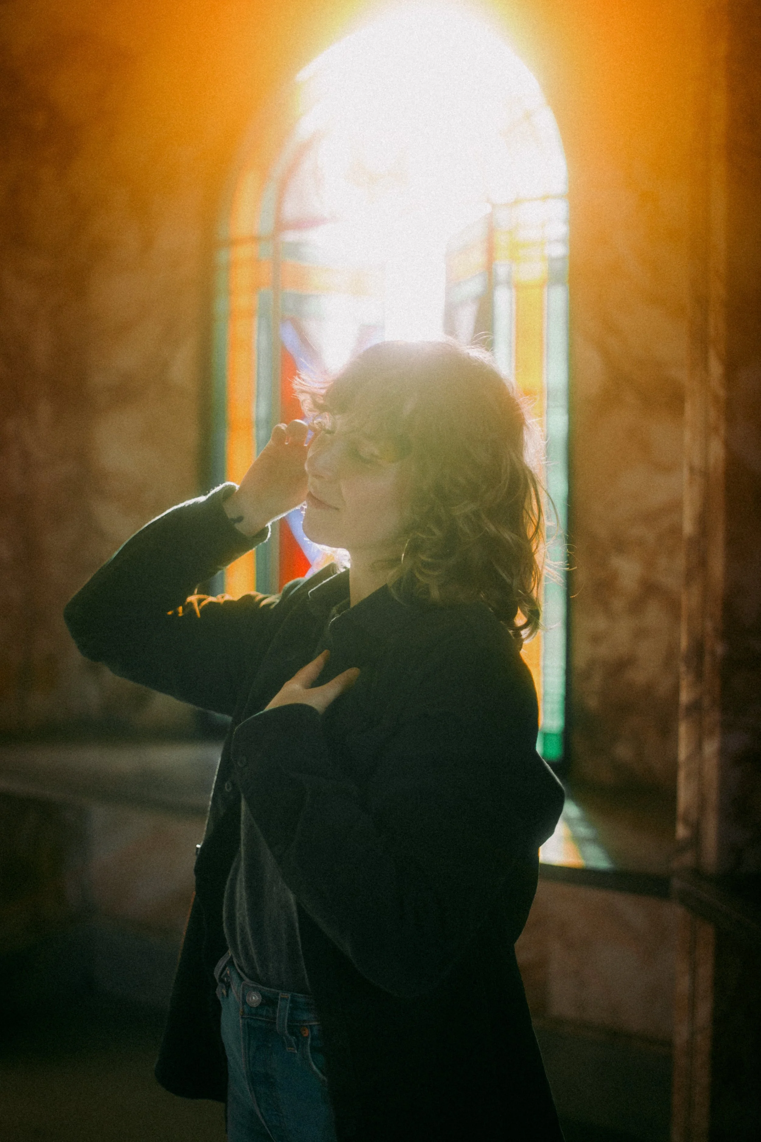Alisha stands in front of a stained glass window. Light pours in behind her, casting beautiful colours. Her wavy hair and a gentle smile is silhouetted in the light. She has raised one hand as though to caress her own cheek and bathe in the light.