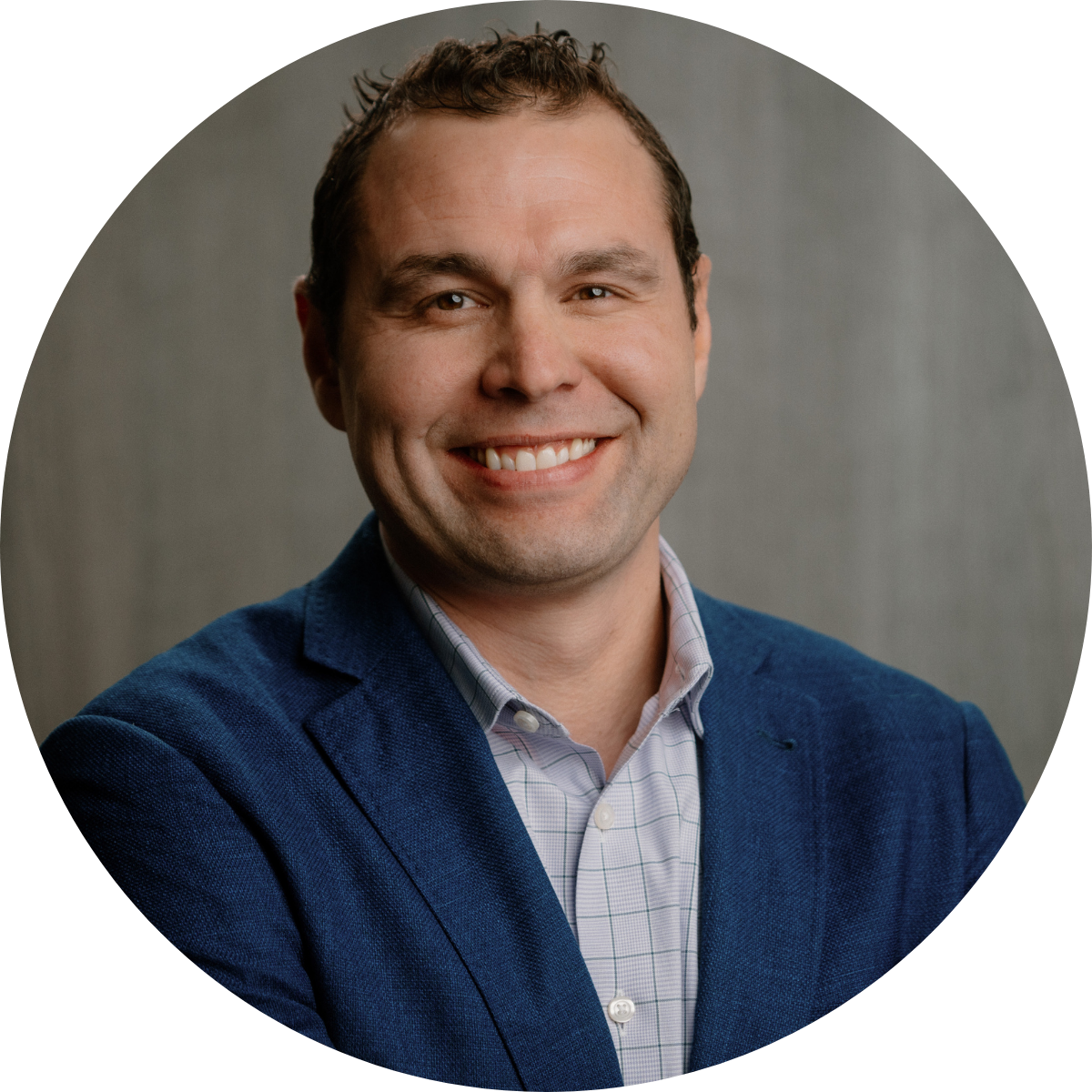 A professional headshot of a smiling man with short, curly hair, wearing a blue blazer and a light-colored checkered shirt, against a plain gray background.