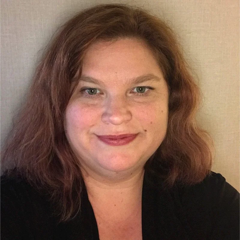 A woman with wavy, shoulder-length reddish-brown hair, light-colored eyes, and fair skin smiling at the camera, wearing a black top against a plain neutral background.