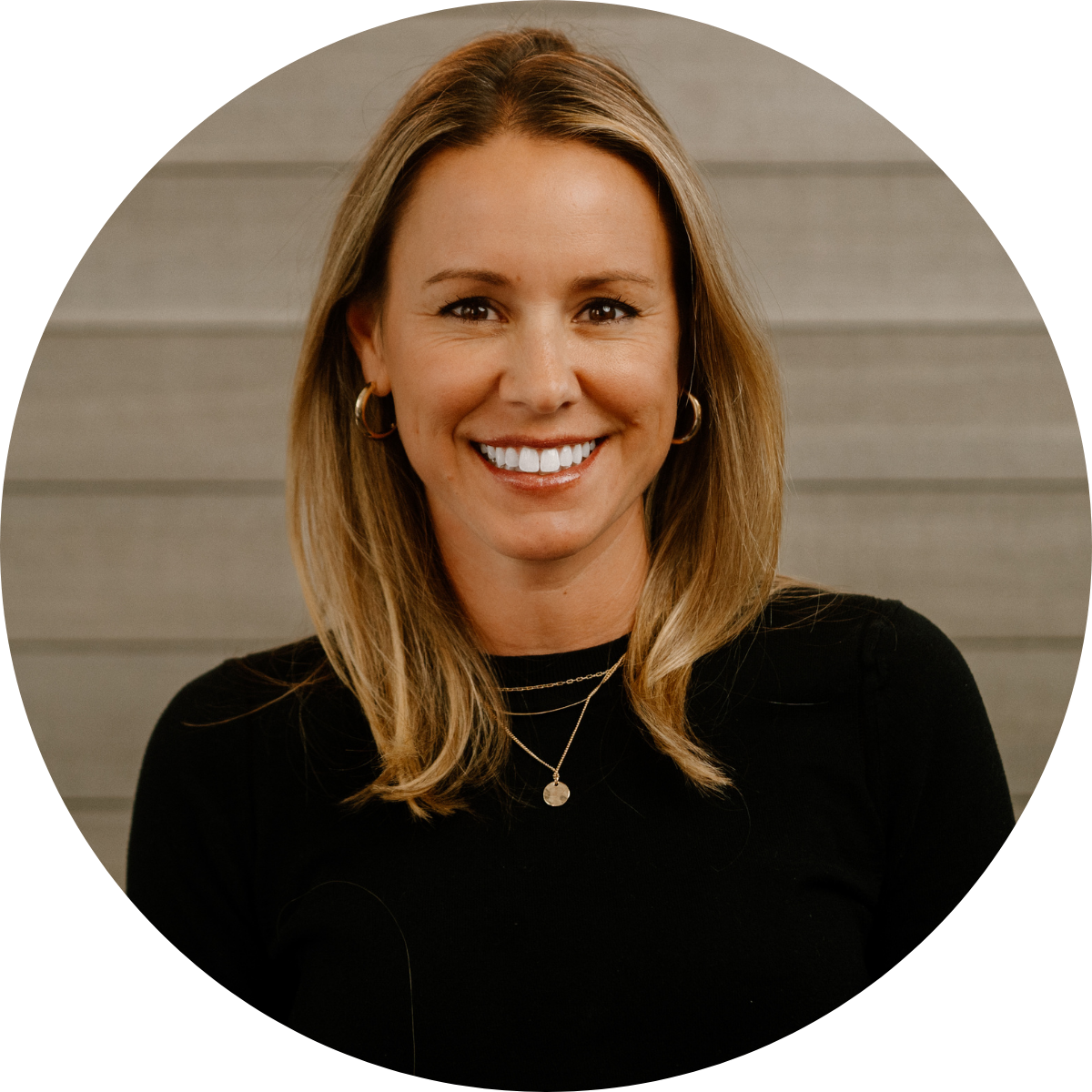 A woman with blonde hair smiling, wearing gold hoop earrings, layered necklaces, and a black top, standing against a wooden wall background.