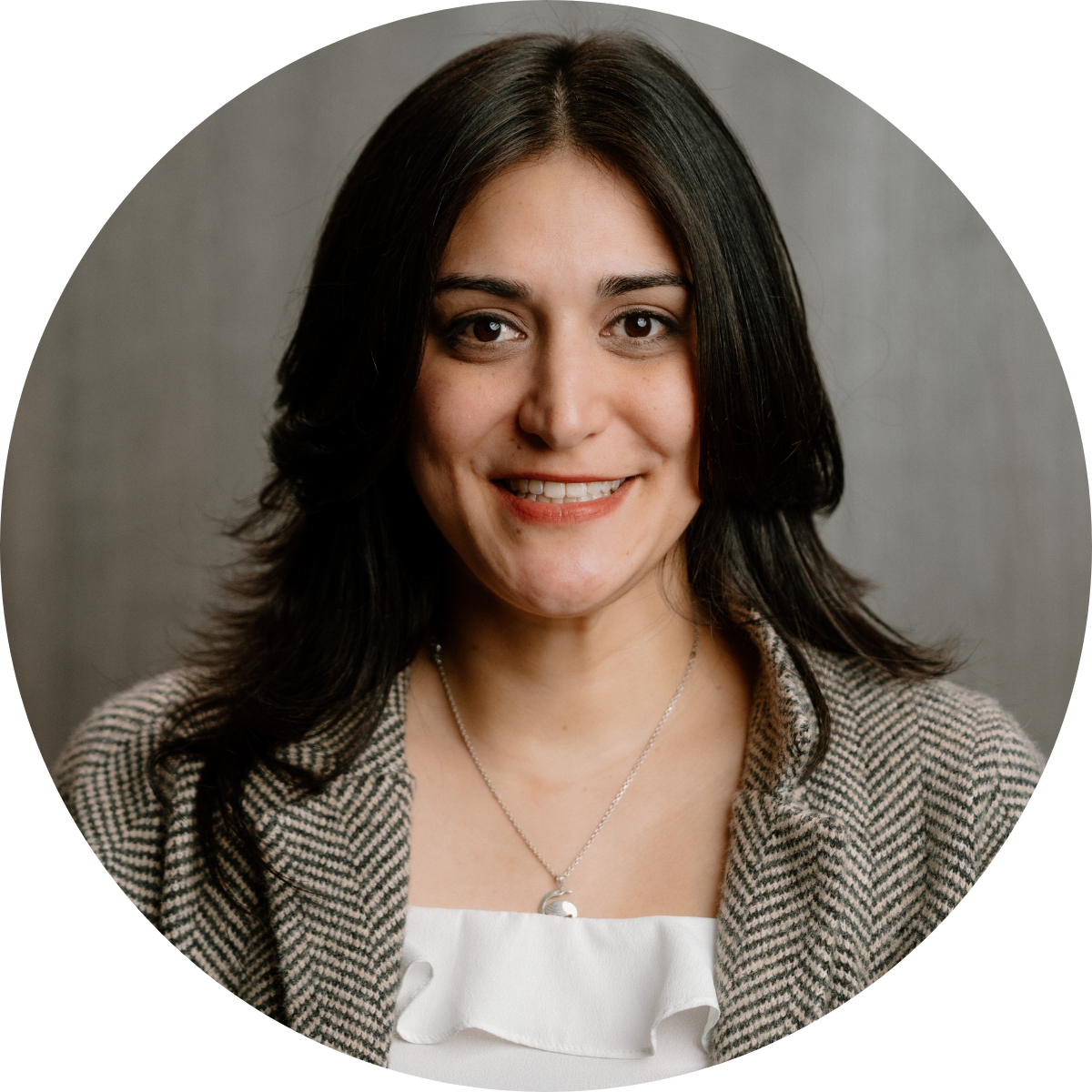 A woman with long dark hair, smiling, wearing a checkered blazer and a white top with a necklace, against a gray background.