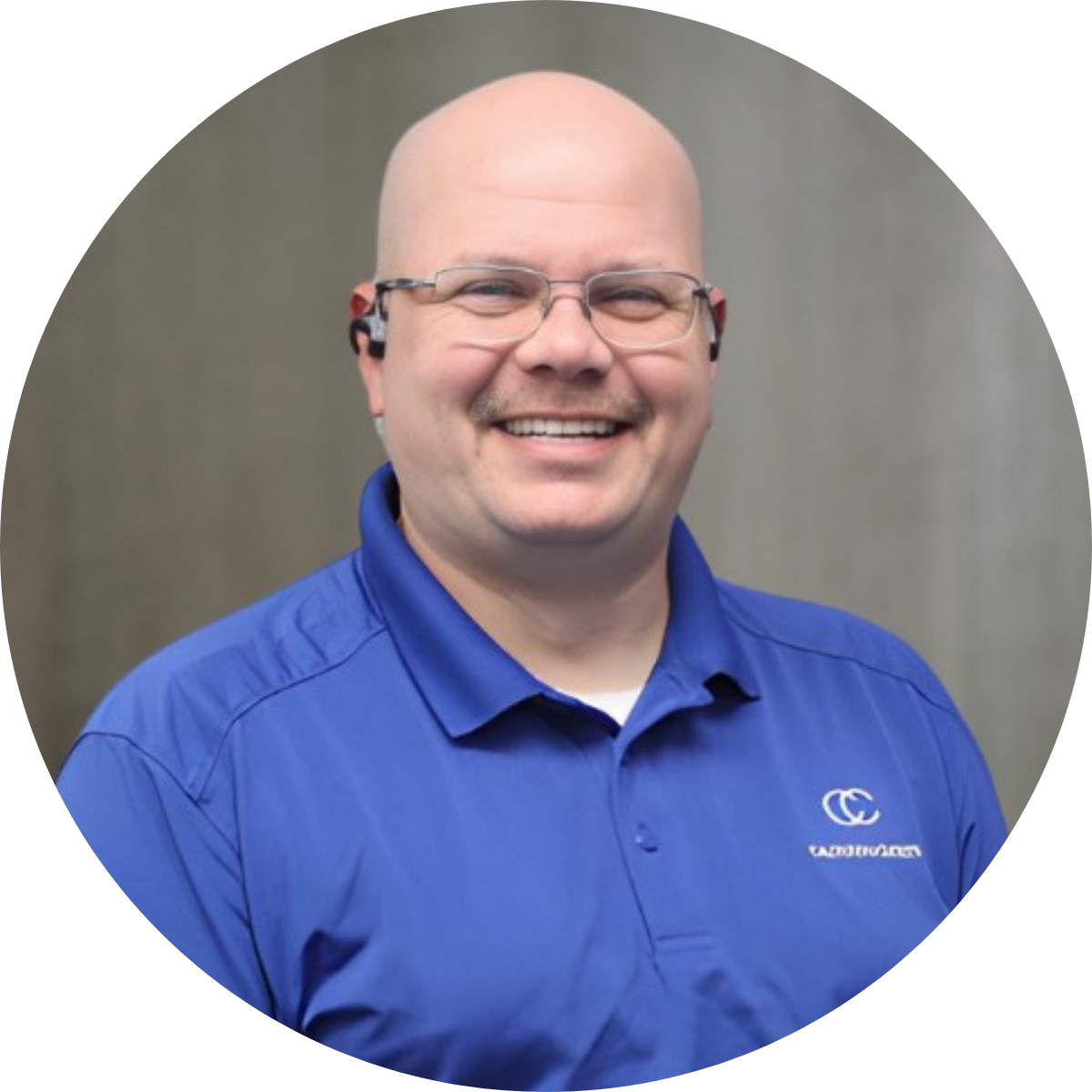 A smiling man with glasses and a bald head, wearing a blue collared shirt with a logo on it, standing in front of a neutral background.