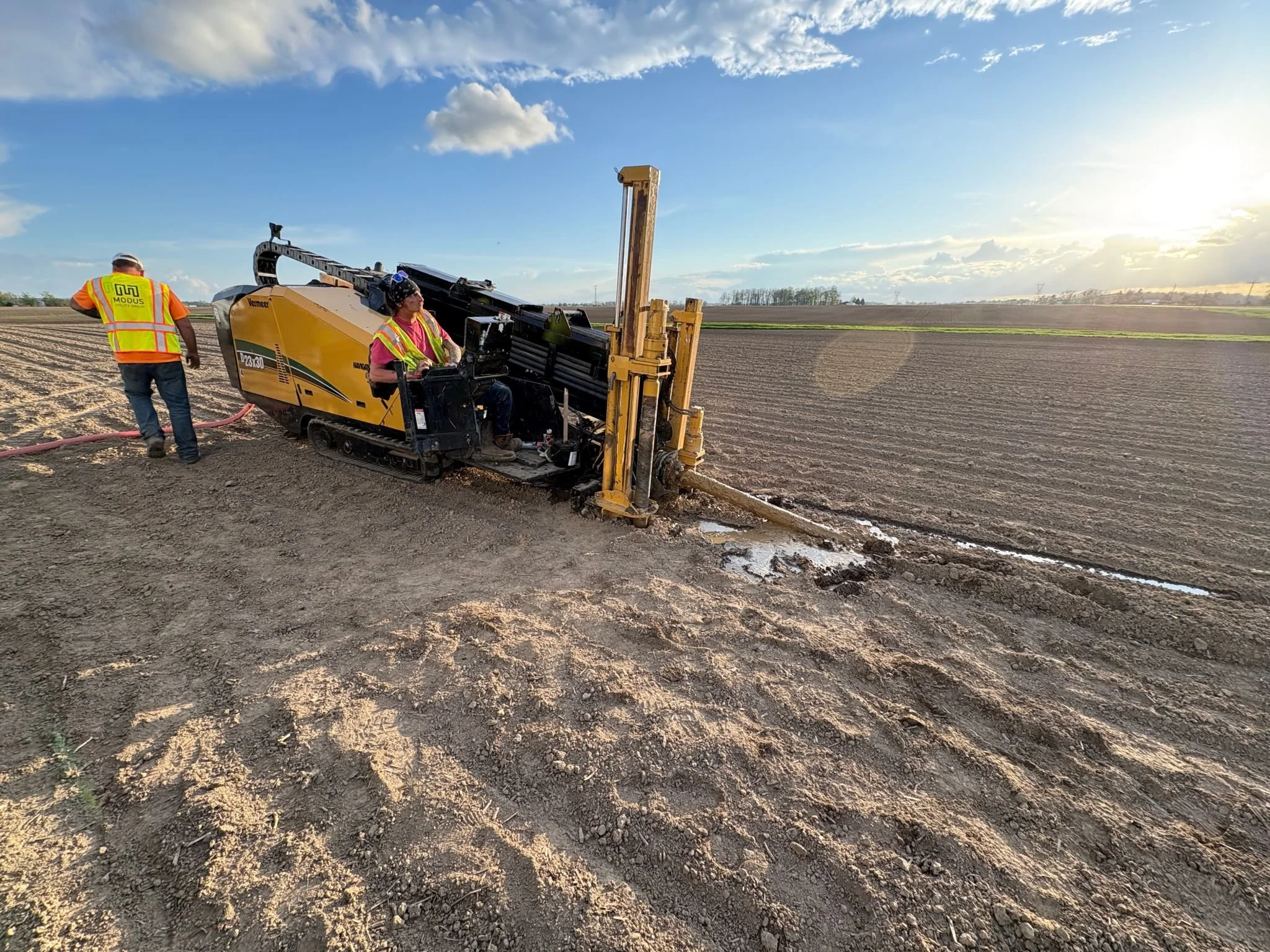 Two workers in orange and pink safety vests operate a yellow ditch digging machine on a farm field during sunset.