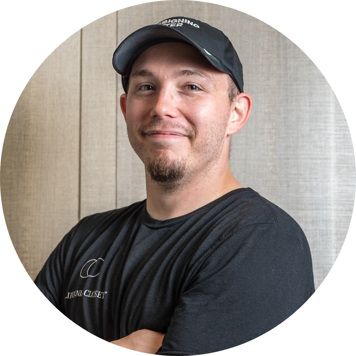 A smiling man with a beard and goatee wearing a black cap and a black T-shirt with crossed arms, standing in front of a beige wooden wall.