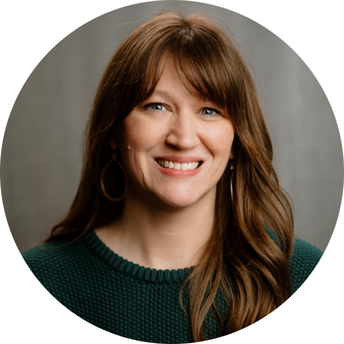 A woman with long, wavy brown hair and blue eyes smiling, wearing a green knit top and hoop earrings, against a gray background.