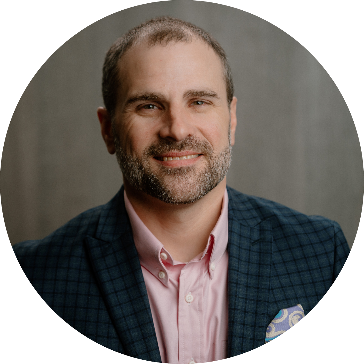 A middle-aged man with short brown hair and a beard, wearing a pink shirt and a dark plaid blazer, smiling in front of a plain gray background.