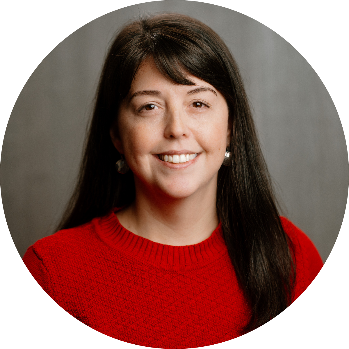 A woman with long dark hair wearing a red sweater and earrings, smiling against a gray background.