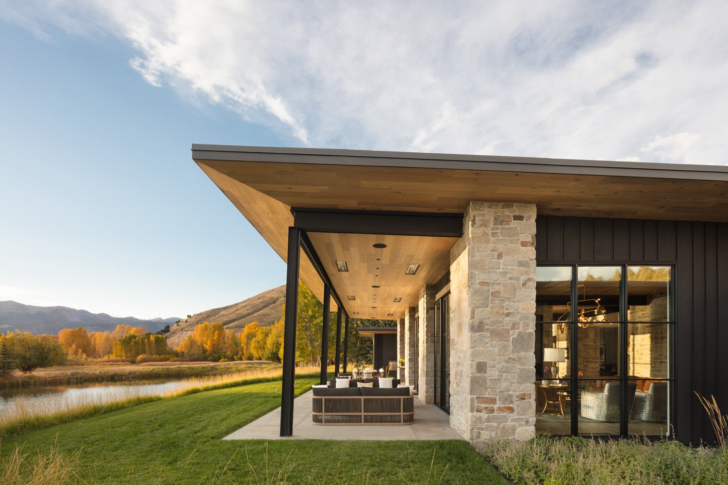 Modern house with stone and wood exterior, large windows, and a covered patio overlooking a scenic landscape with mountains, grass, and autumn trees by a lake.
