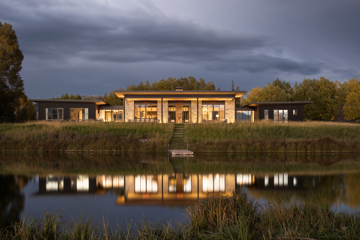 Modern house with large windows reflected in a calm lake, surrounded by trees and grass under a cloudy sky.