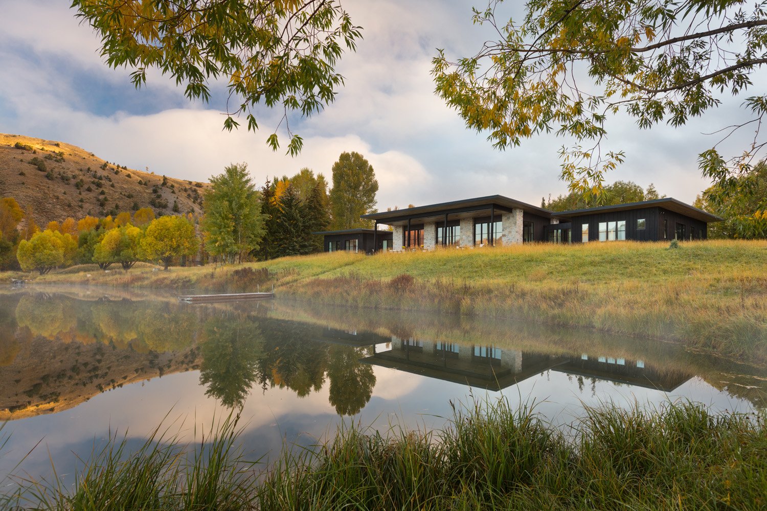 Modern lakeside house with large windows in a scenic landscape of trees, grassy hills, and a reflective pond. Autumn foliage is visible.