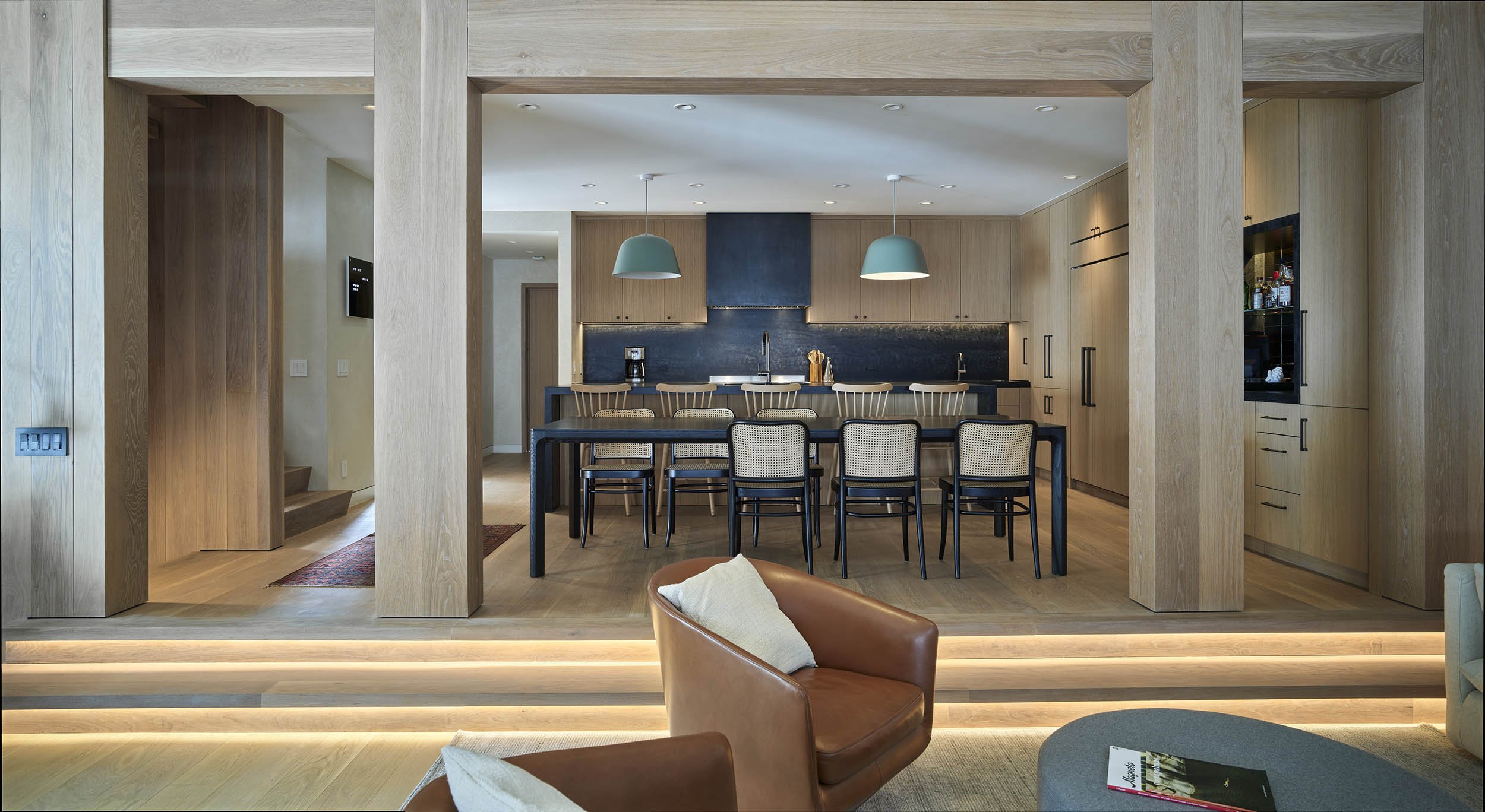 Modern kitchen and dining area with wooden cabinets, a long dining table with wicker chairs, pendant lights, and a cozy seating area with leather armchairs and a coffee table.