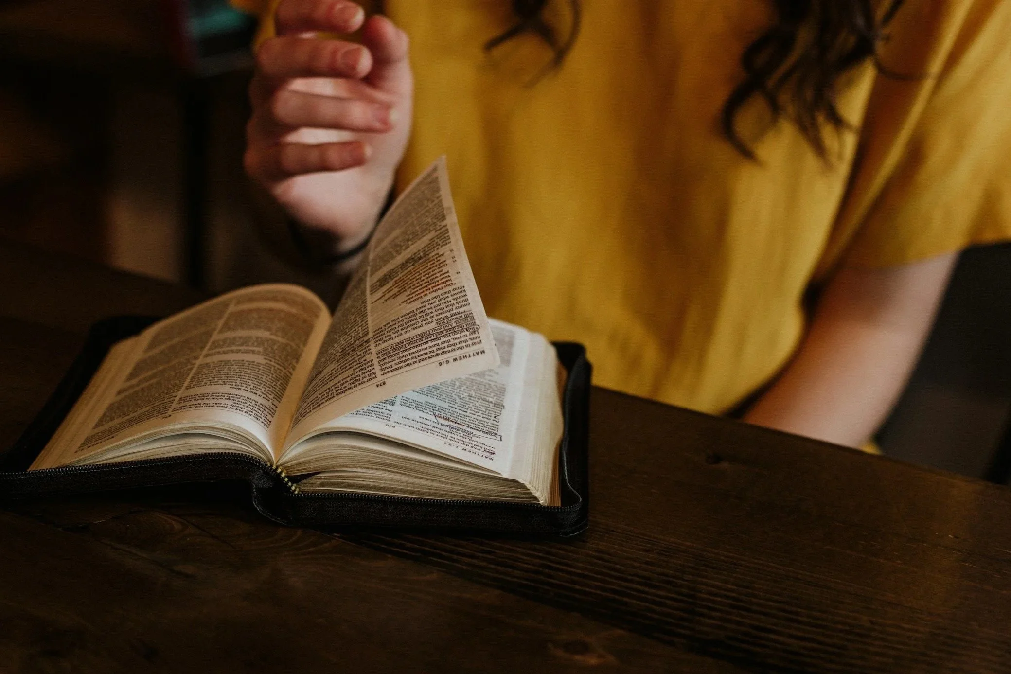 A person in a yellow shirt reading a book with a black cover, placed on a wooden table.