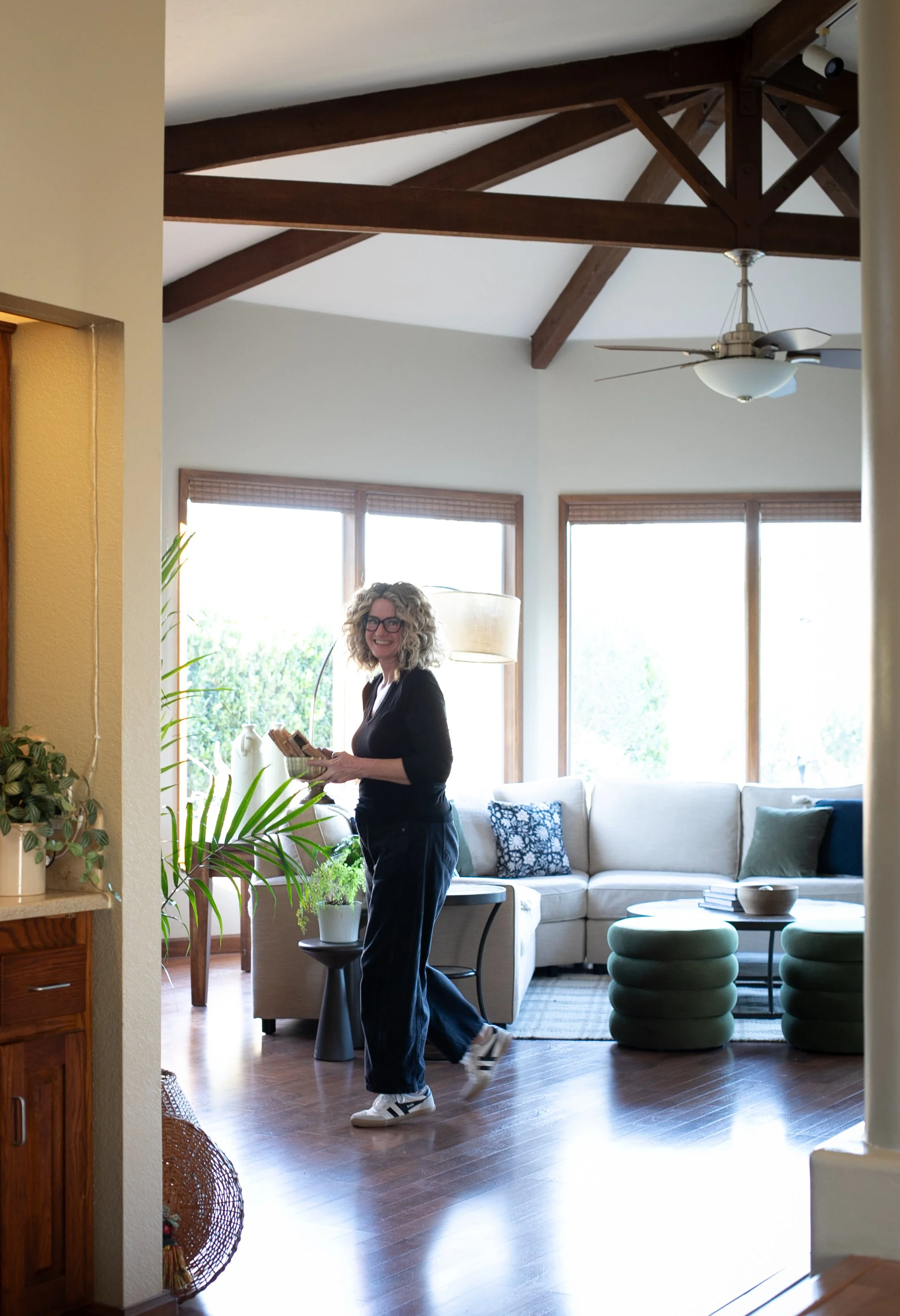 A smiling woman with curly blonde hair, glasses, wearing a black long-sleeve shirt, black pants, and white sneakers, standing in a bright living room holding a tray of food. The room features large windows, a white sofa with decorative pillows, indoo