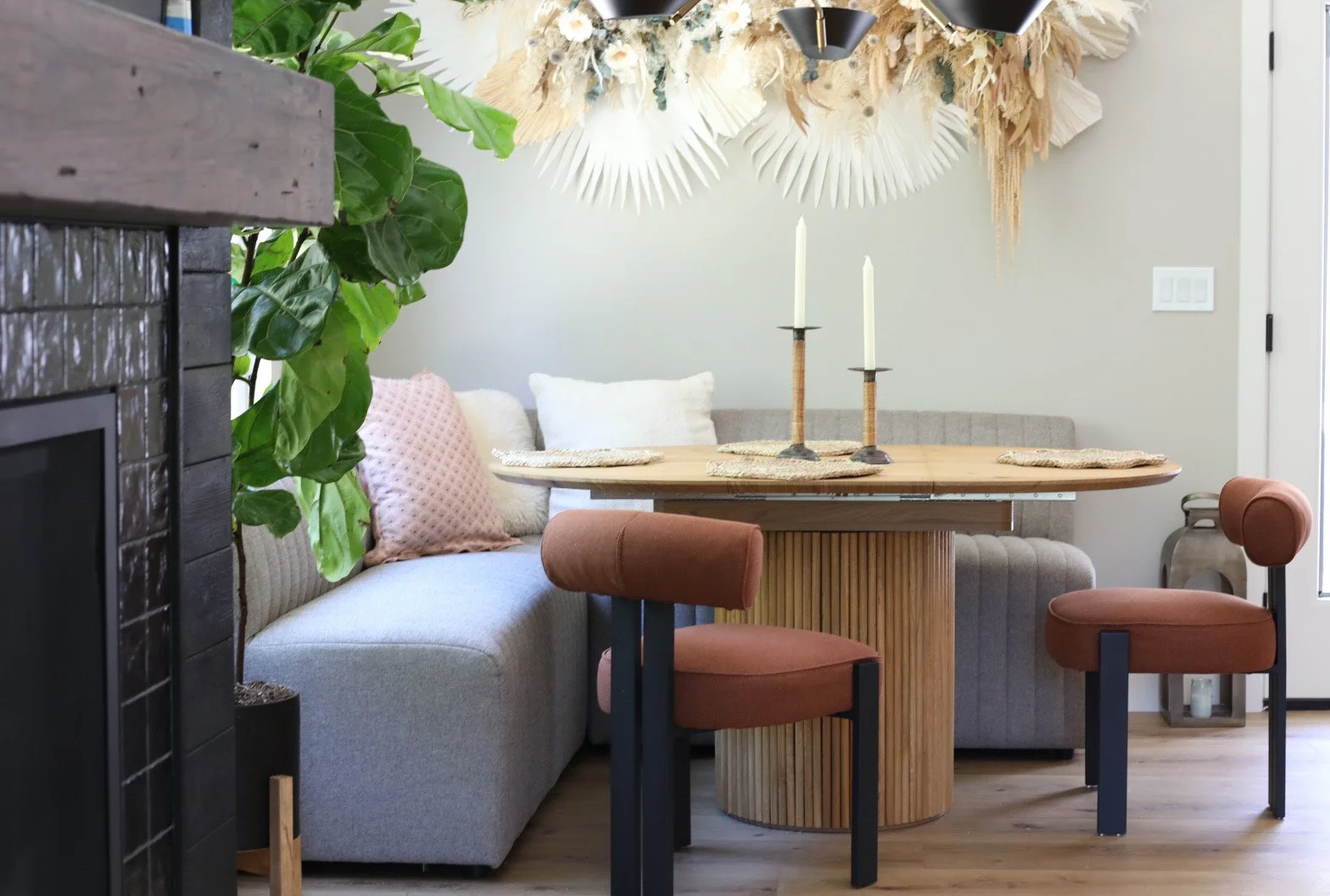 A modern dining room with a light gray sofa and pink pillows on the left, and a round wooden table with two rust-colored chairs on the right. The table has two candlesticks with white candles. A decorative wall hanging made of dried leaves and flower