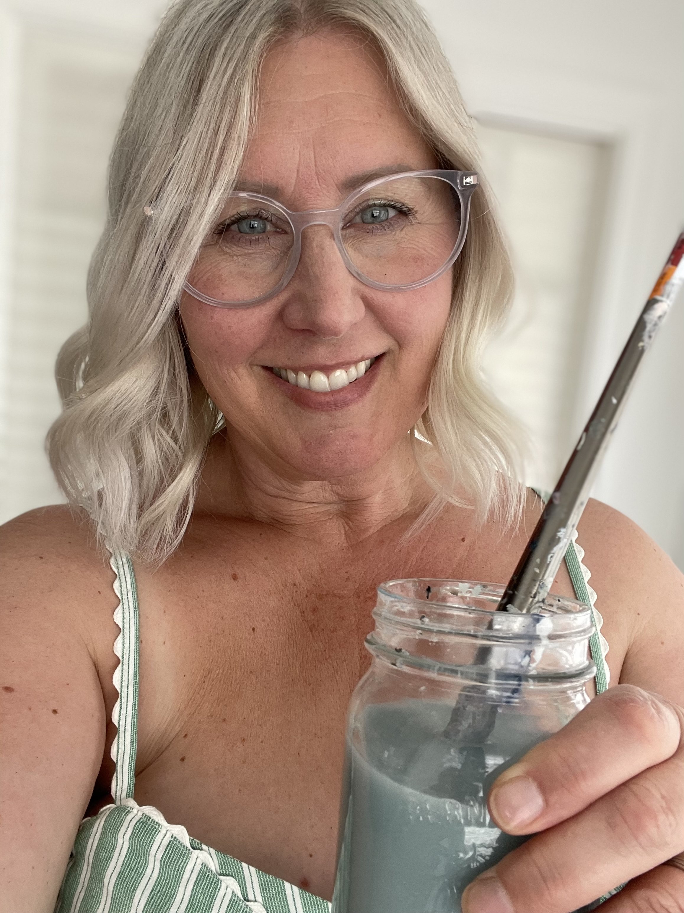 Smiling woman artist Cathy Empey with blonde hair and clear frame glasses holding a mason jar of blue paint with a paintbrush as she paints abstract Love Letters to the Lakes of the Okanagan.