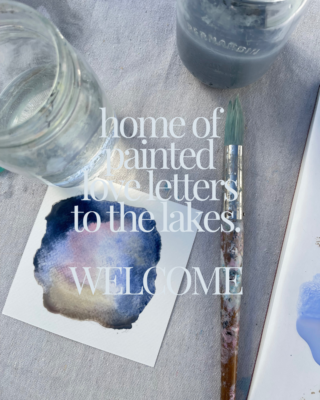 A watercolor painting with shades of blue, purple, and beige on paper, surrounded by two jars of water, an artist's brush with paint on the handle, and overlaid white text that reads, "home of painted love letters to the lakes. WELCOME."