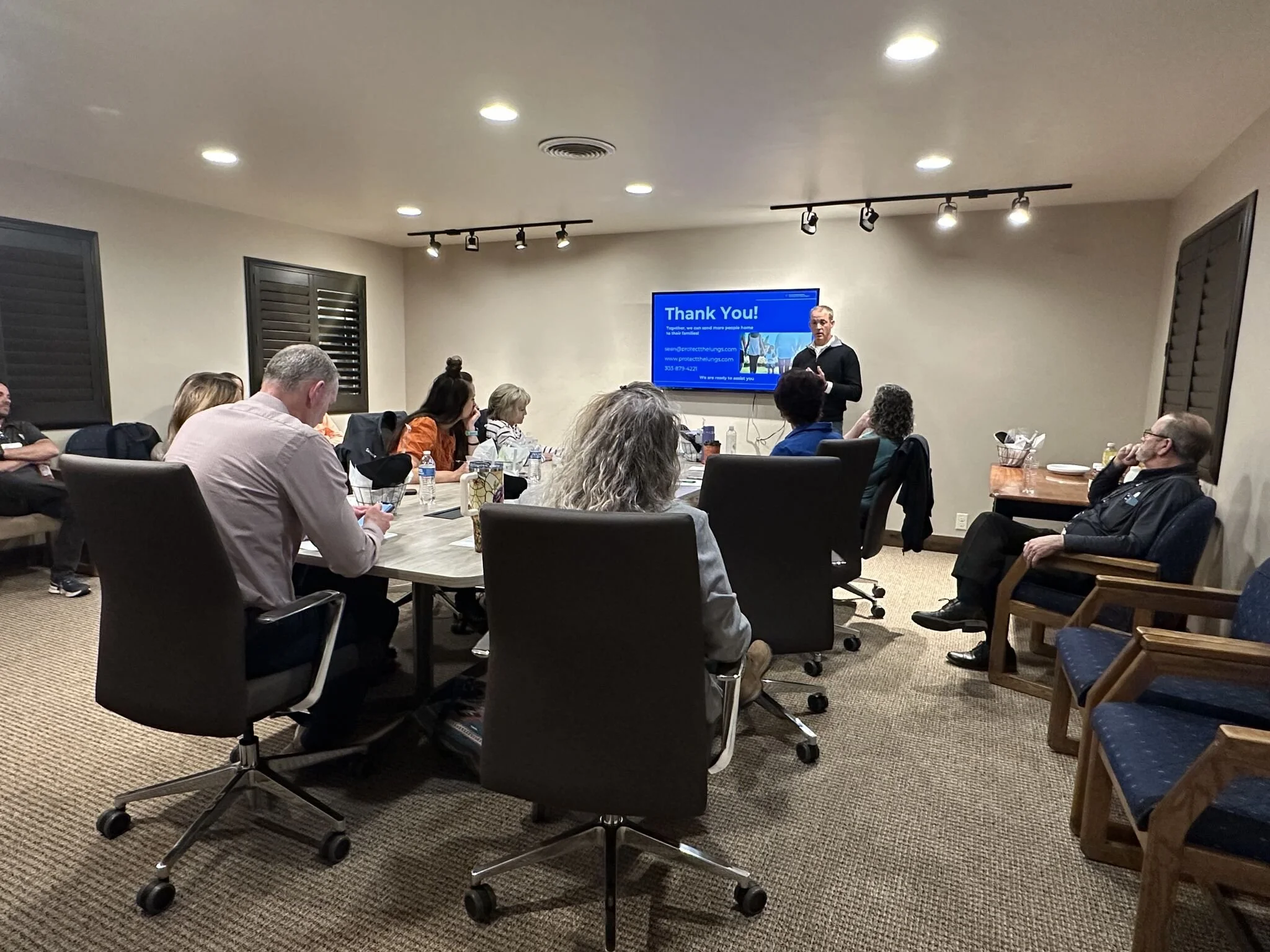 A group of people in a conference room attending a presentation. The presenter is standing near a blue screen with a