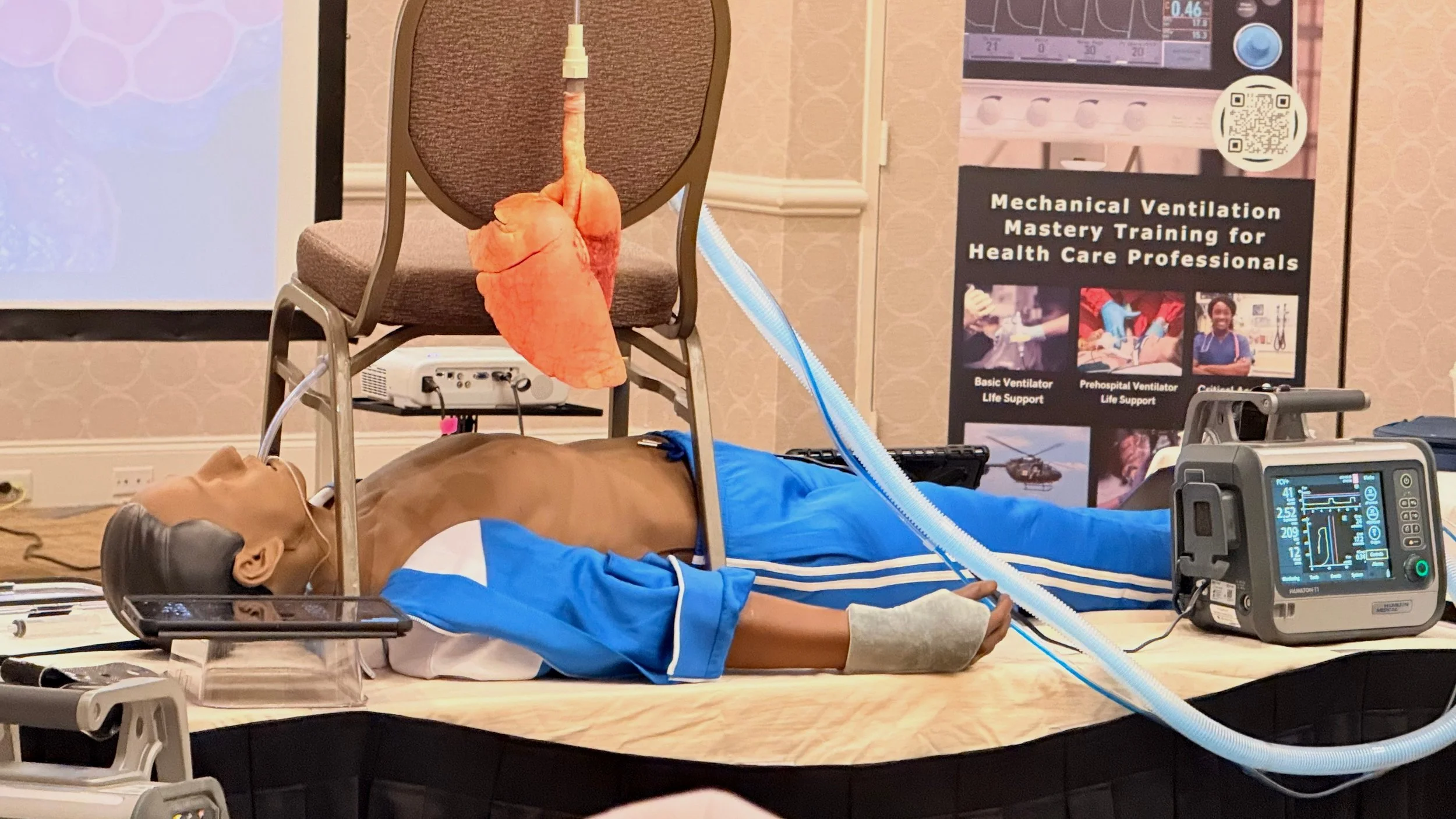 Medical training mannequin lying on a table connected to ventilator equipment, with a model of human lungs hanging from a chair, in front of a banner about mechanical ventilation training for health care professionals.