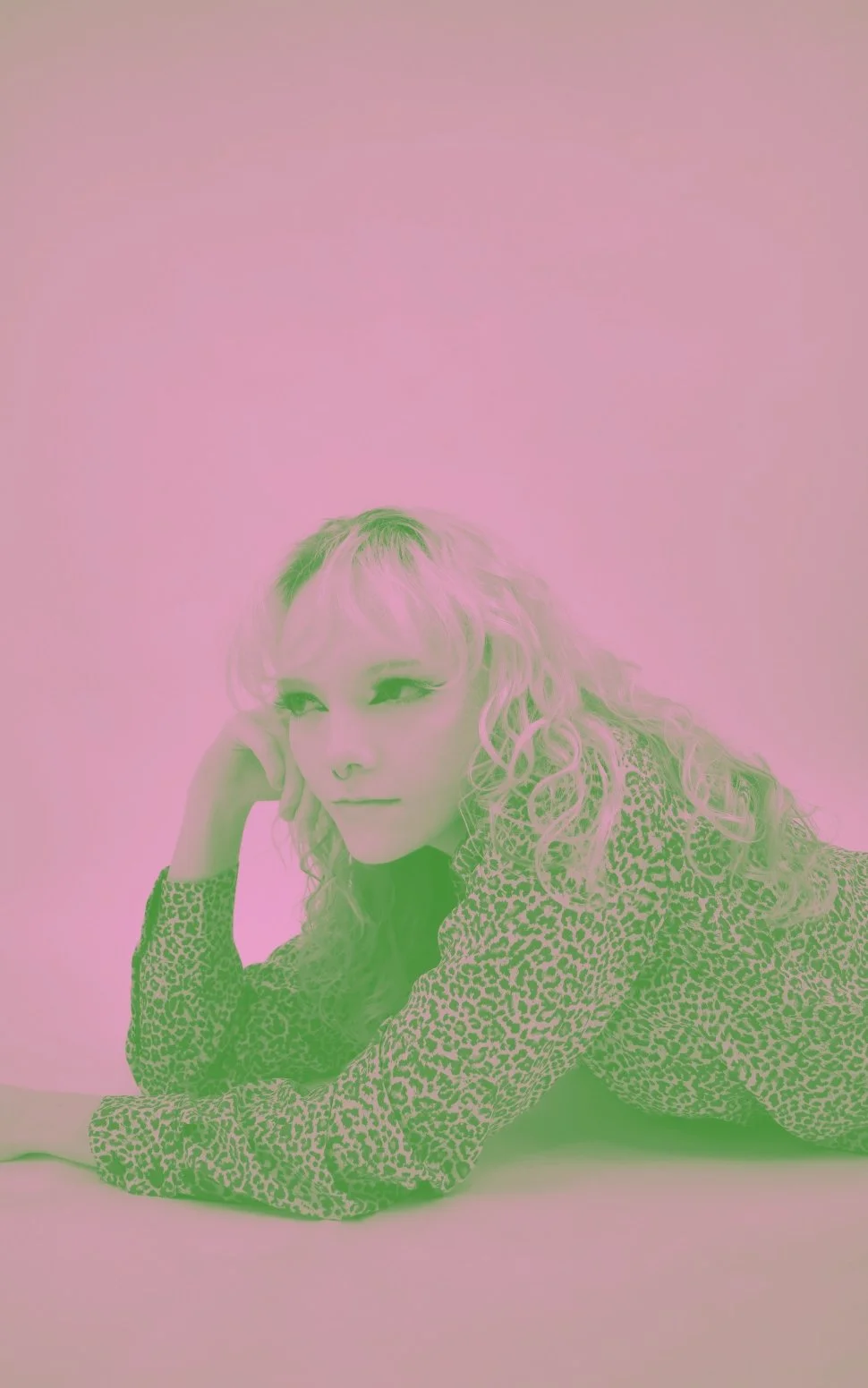 A woman with curly hair lying on her side against a pink background, wearing a leopard print top.